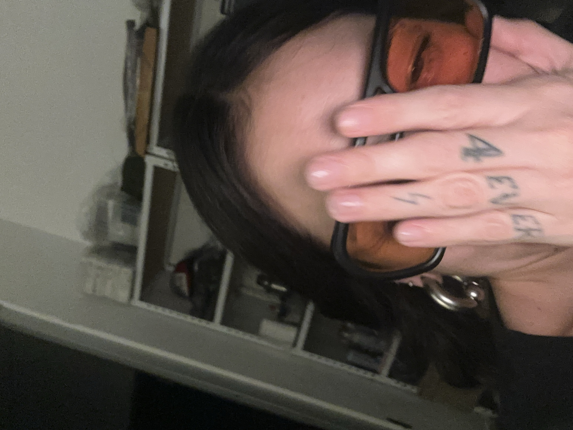 The image presents a close-up, dimly lit indoor scene. A person with dark hair, parted centrally, is partially depicted. Their right hand is raised, covering the left side of their face, including the forehead and the area around the left eye. The individual wears black-framed glasses with orange-tinted lenses; the right eye is partially visible through its lens. A silver-colored hoop earring or tunnel is visible in the left ear. Several tattoos are present on the fingers of the right hand: a small lightning bolt on the middle finger, the inverted text "EVER" on the ring finger, and the inverted numeral "4" on the pinky finger. The background is an out-of-focus, light-colored shelving unit displaying various indistinct objects. The specific location within the city of Buenos Aires, Argentina, cannot be determined from the visual information provided in the image.