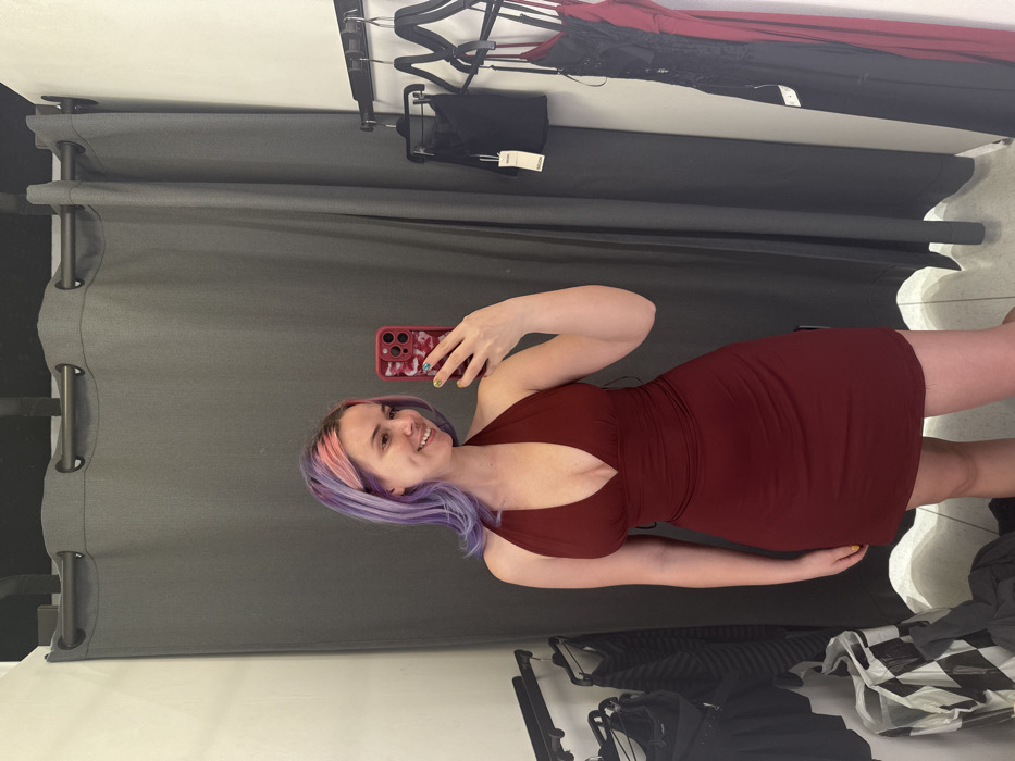 In a retail changing room in the City of Zagreb, Croatia, a female subject is depicted from the waist up, holding a red smartphone in her right hand to capture a mirror selfie. The female, appearing in her late teens or early twenties, has long, wavy hair with purple and pink sections, a light complexion, and a smiling expression. She is wearing a dark red, halter-neck mini-dress featuring a deep V-neckline and gathered fabric at the waist. Her fingernails are painted with a multi-colored design on some and yellow on others. The background features dark grey privacy curtains hung from a black metal rod. To the left, a metal rack holds various dark garments, and a black and white checkered shopping bag rests on the floor. To the right, a second metal rack displays additional clothing, including a black lace dress and a dark red dress. The floor is light-colored, possibly tiled.
