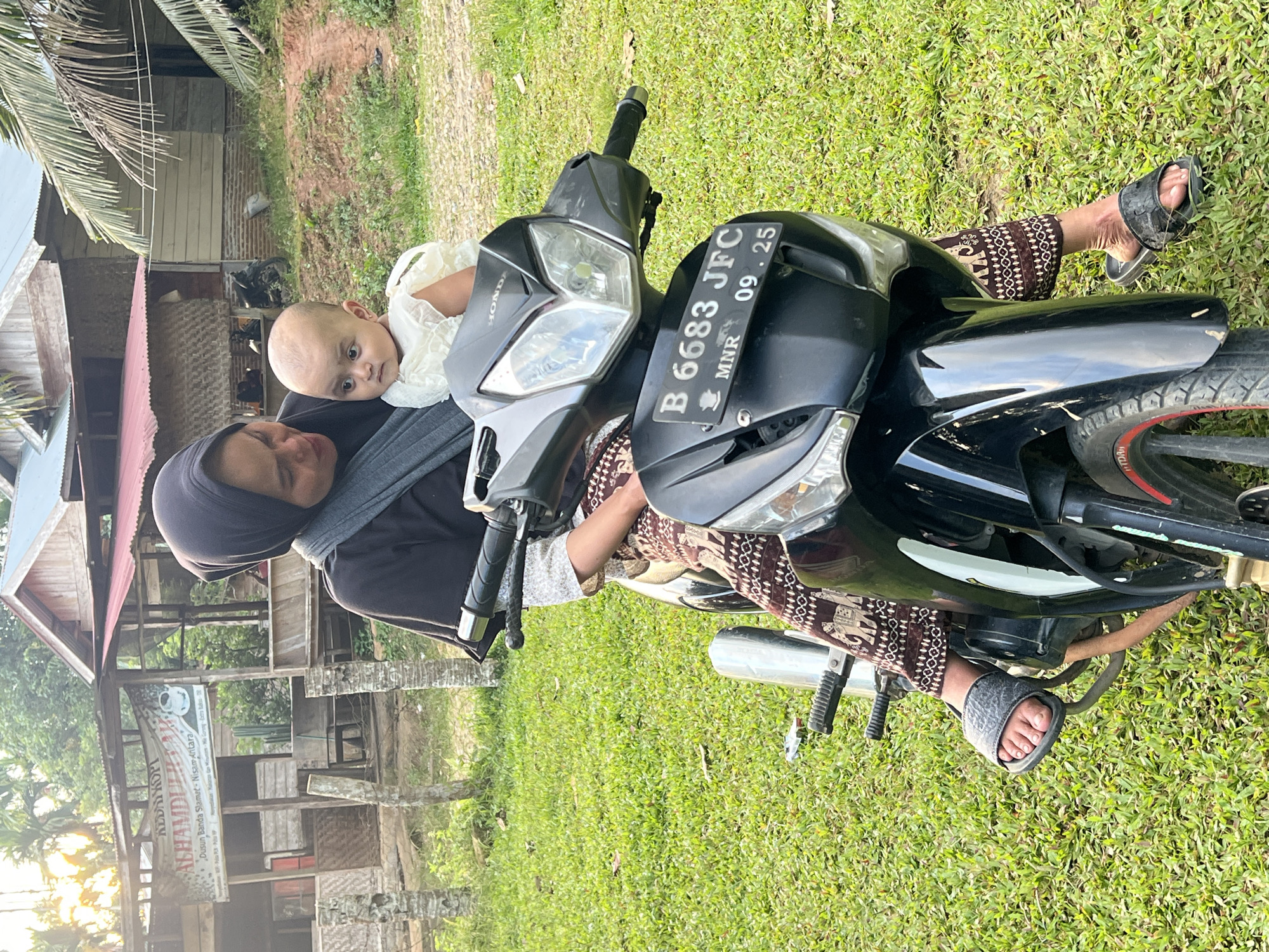 In Kilometer Delapan, Indonesia, a woman wearing a black hijab and a patterned brown skirt sits on a black motorcycle. She is holding a baby who is wearing a white dress. The woman appears to be looking down at the baby, and the baby is looking forward. They are on a grassy area with a building in the background. The building has a red roof and is made of wood. There is a sign on the building that reads "ALHAMDULILLAH KOPI". The license plate on the motorcycle reads "B 6683 JFC MNR 09.25". The lighting suggests it is daytime, possibly late afternoon given the warm tones of the light.