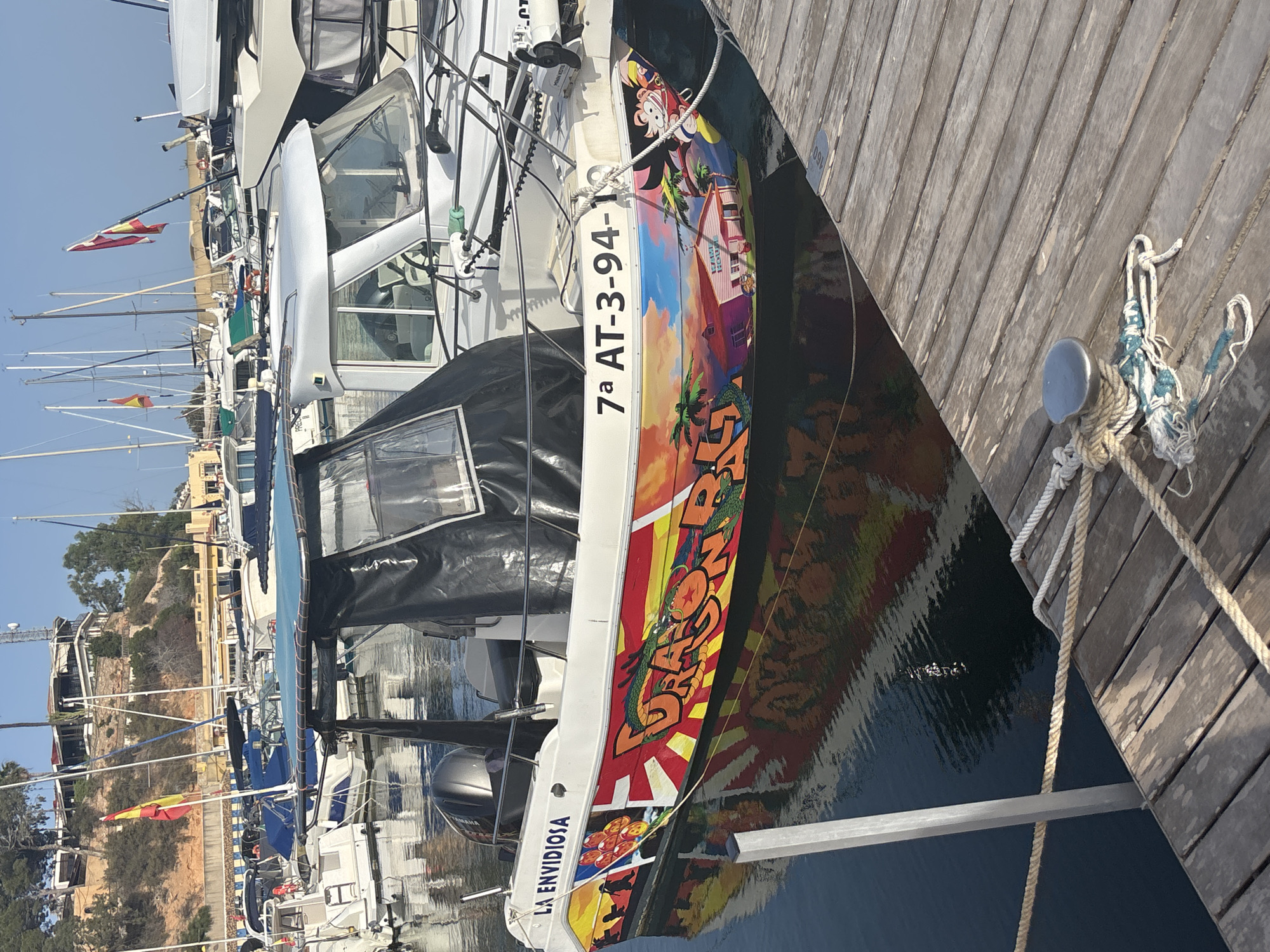 A white motorboat named "LA ENVIDIOSA" is docked at a wooden pier in a marina. The boat has a colorful mural on its side, featuring characters from the Dragon Ball franchise, palm trees, and a house. The boat's registration number, "7a AT-3-94-19", is visible on its hull. Other boats and masts of sailboats are present in the background. The water is calm and reflects the boat and the surrounding environment. The sky is clear and blue. The overall scene depicts a sunny day at a marina.