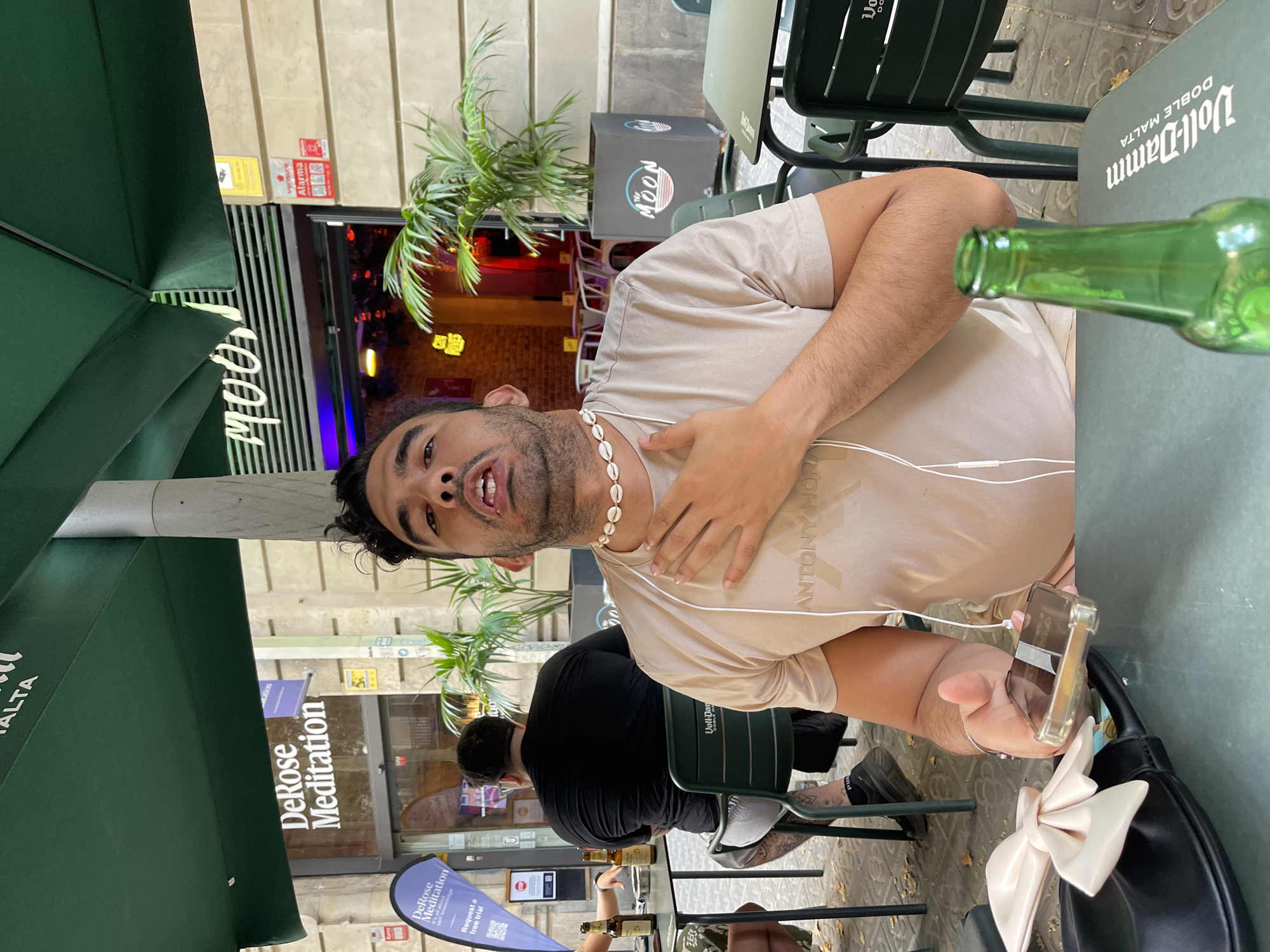The image shows a man in Barcelona, Spain, seated at an outdoor table in front of a cafe. He is wearing a tan t-shirt and a seashell necklace. His right hand is on his chest, and he is holding a phone in his left hand. A green bottle of Voll-Damm beer is in the foreground, and a black purse with a large white bow is on the table. In the background, there is a sign that reads "DeRose Meditation" and another sign with "MOON" written on it. Other people are visible at tables in the background, and palm plants are present. The scene suggests a casual outdoor dining or drinking setting.