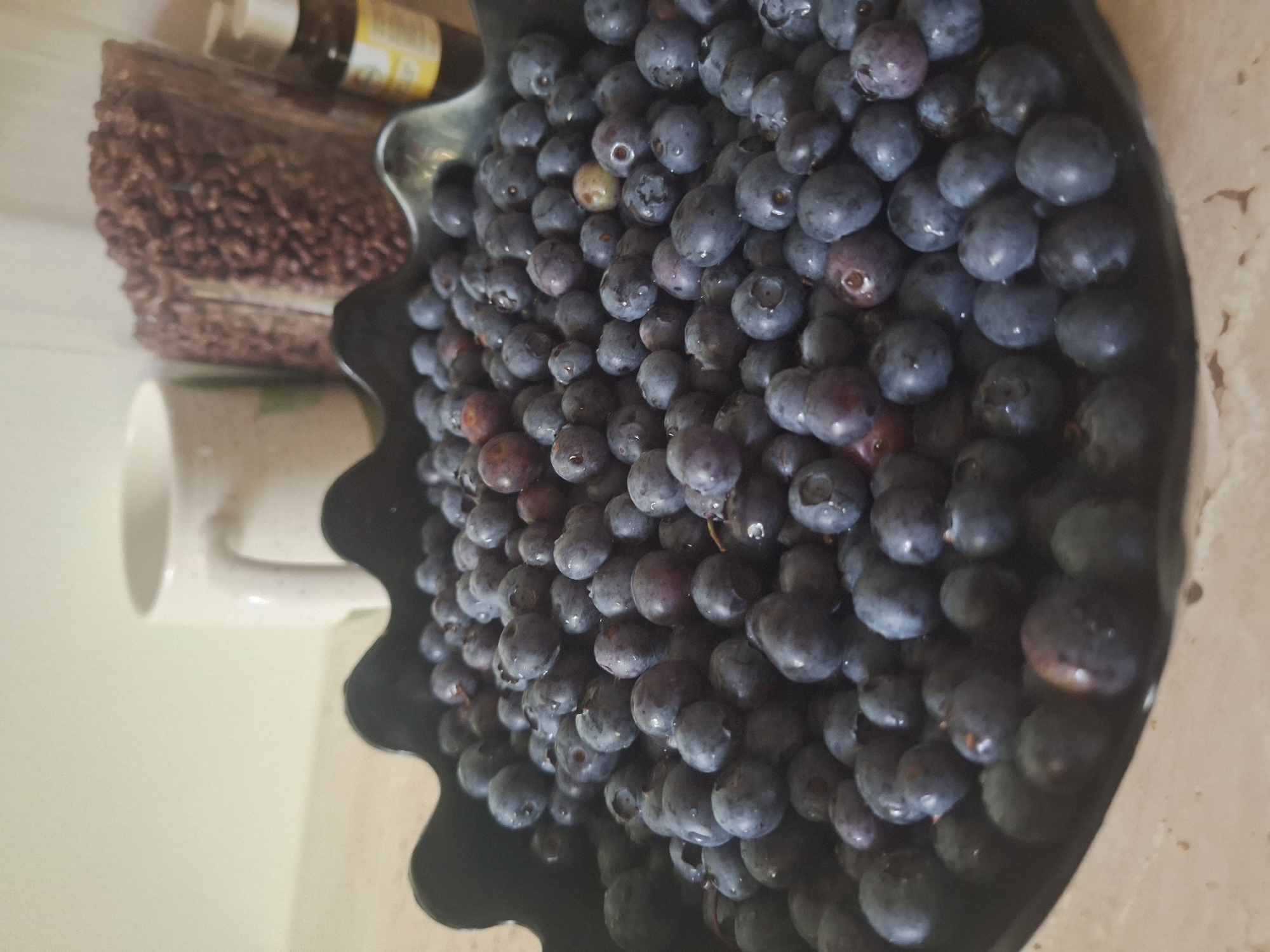 A close-up, slightly angled shot shows a dark, wavy-rimmed bowl overflowing with fresh blueberries. The blueberries are a deep blue-purple, some with a matte finish and others glistening with moisture. Behind the bowl, to the left, is a speckled white ceramic mug. To the right and slightly behind the mug is a tall, clear glass container filled with small, dark, granular items, possibly dried beans or cereal. Next to the glass container is a small, amber-colored glass bottle with a white dropper lid. The background is a plain, light-colored wall. The scene is indoors, with the objects resting on a light-colored surface. No specific location within Mińsk Mazowiecki, Poland can be identified from the image.