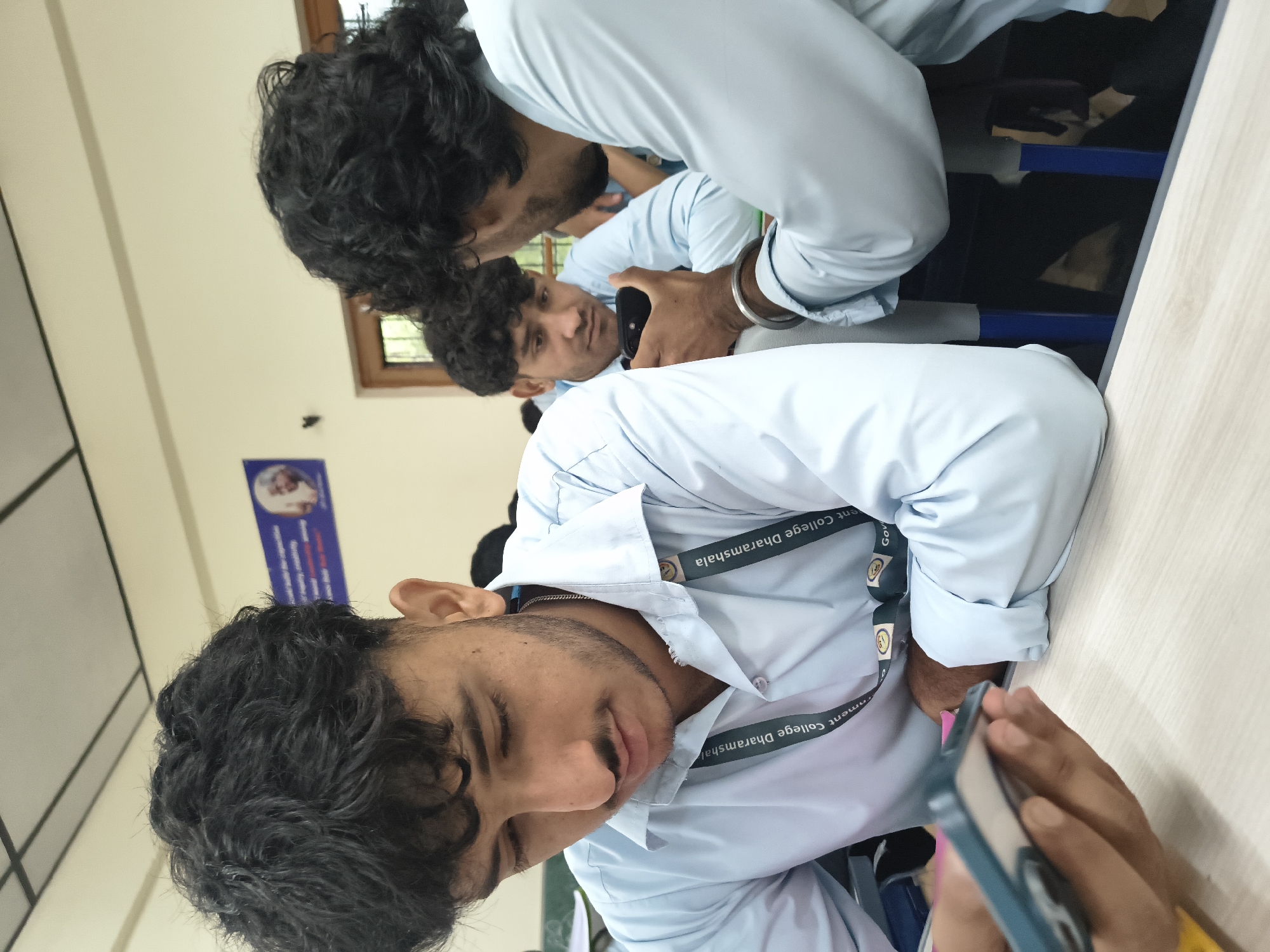 In a classroom setting in Dharamshala, India, three young men in light blue collared shirts are seated at a wooden table, each engaged with their mobile phones. The person in the foreground is looking down at a blue-cased phone held in their left hand, while a silver chain is visible around their neck. To their right, another individual in a light blue shirt is showing something on a black mobile phone to a third person, who is also looking at the phone. Both of these individuals have dark, curly hair. A lanyard with text reading "Government College Dharamshala" is visible around the neck of the person in the foreground. The classroom has a beige ceiling with visible fluorescent lighting fixtures. In the background, a blue poster with an image of a person is affixed to the wall. The environment suggests an educational institution, likely a college campus in Dharamshala.