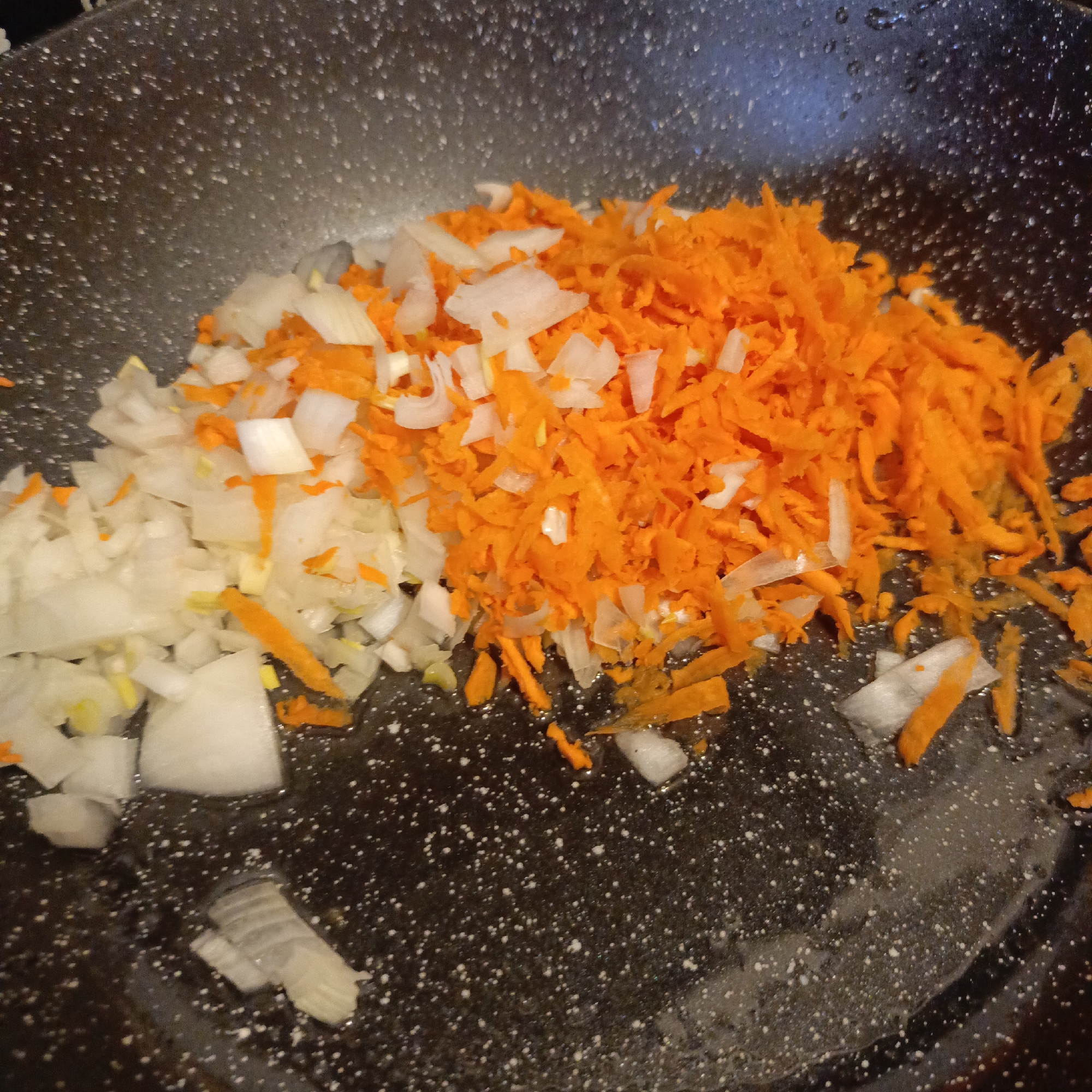An overhead close-up view captures a dark-colored cooking pan filled with two types of raw, chopped vegetables. On the left, there is a pile of diced white onions. To the right, a larger quantity of shredded bright orange carrots is visible. The onions and carrots are partially mixed in the central area of the pan. The pan itself has a speckled texture, characteristic of some non-stick cookware. There appears to be some liquid, likely cooking oil, in the bottom of the pan, reflecting light in various spots. The scene suggests food preparation, with the vegetables ready to be cooked, presumably in a kitchen. There is no visible text or indication of the time of day.