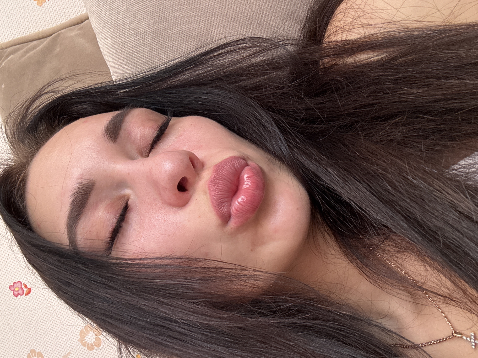 A close-up, high-angle shot shows a woman with dark hair and closed eyes, lying down. Her lips are slightly puckered. The background consists of neutral-toned cushions with a floral-patterned surface visible on the left side of the frame. A delicate gold necklace with a cross pendant is visible on her neck. There's no indication of Chaikovsky, Russia.
