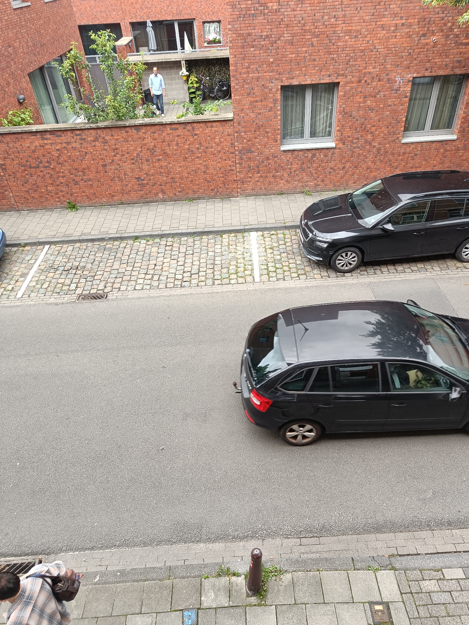 The image presents an elevated view of a street scene in Leuven, Belgium. The scene is dominated by red-brown brick buildings and a street with parked vehicles and pedestrians.
In the upper left, a multi-story brick building features a balcony or terrace area enclosed by a high brick wall at street level. A male individual stands on this balcony, clad in a light blue long-sleeved shirt, dark trousers, and white shoes. He faces generally towards the left. The balcony also hosts several green plants, a black cylindrical bin, a black bicycle parked against a patterned screen, a folded white parasol or canopy, and a planter containing yellow flowers. To the right, another red-brown brick building displays two rectangular windows, each fitted with grey frames and light-colored interior curtains.
The street below comprises an asphalt road and a dual-sectioned sidewalk. The section immediately adjacent to the asphalt is paved with irregular, light-toned cobblestones, where a rectangular metal drain cover is visible. A white line painted on the cobblestones delineates a parking space. A black Skoda SUV or station wagon, consistent with a Kamiq or Scala model, is parked in this section, facing left. The second sidewalk section, closer to the viewer's vantage point, is composed of large, light grey square paving slabs.
On the asphalt road, a second black Skoda station wagon, identifiable as an Octavia model, is positioned, also facing left. Its glossy roof reflects the sky and portions of unseen foliage. A tow hitch is present at the rear of this vehicle.
In the lower left foreground, a second individual is partially visible, seen from the shoulders up. This person wears a light-colored plaid shirt and carries a dark brown shoulder bag, with their head angled downwards. Sparse green weeds grow from cracks in both the cobblestone and paved sidewalk sections, as well as along the base of the brick walls. A dark brown bollard is situated on the paved sidewalk in the lower right portion of the frame.