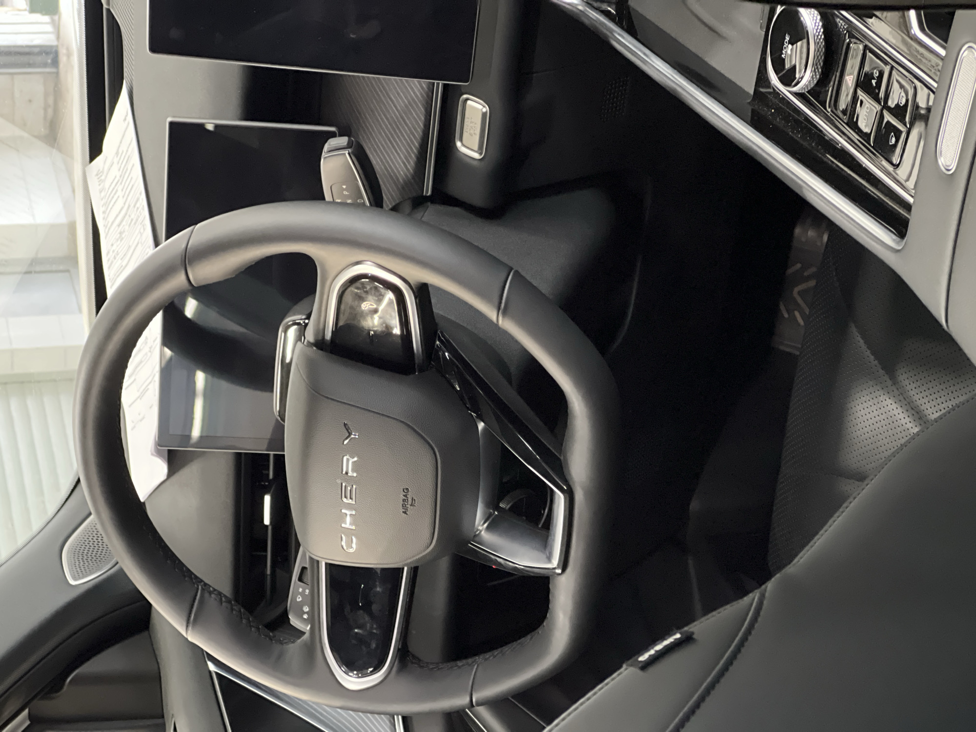 The image captures the interior of a modern car, likely from the driver's perspective, focusing on the steering wheel and central console. The steering wheel is black, leather-wrapped, with visible stitching on the rim and a flattened bottom section. The prominent "CHERY" brand name is embossed in silver on the center airbag cover, with the word "AIRBAG" and an associated symbol printed below it. Silver and gloss black trim accents are present on the steering wheel spokes.
Behind the steering wheel, a digital instrument cluster is partially visible, and to its right, a large, dark rectangular screen, presumably part of the infotainment system, is prominent. Another similar screen is visible further to the right. A white document, possibly a vehicle specification sheet or price list, rests on the dashboard, partially obscuring the top of the screens. The dashboard features a textured, dark panel below the windshield and a section with a carbon-fiber-like pattern.
To the right of the steering column, a gear selector stalk displaying "P," "N," and "D" is visible. Below the right-hand display, a small rectangular screen shows "MODE" and "ECO," accompanied by a silver button. Further down and to the right, on the central console, there is a control panel featuring a large, textured rotary knob labeled "MODE" and several push-buttons, including controls for "AUTO," "A/C," hazard lights, and windshield functions. This panel has a glossy black finish with chrome accents.
The driver's seat is upholstered in black leather with visible stitching and perforated sections. A reflective trim piece with a geometric pattern, possibly "AVA," is visible on the lower interior panel or floor near the driver's feet. The overall interior appears clean and well-lit. Through the windshield, reflections suggest an indoor environment, such as a showroom or garage, with light-colored walls and large windows in the background. There are no identifiable visual cues within the image to confirm the specified location of Izhevsk, Russia.