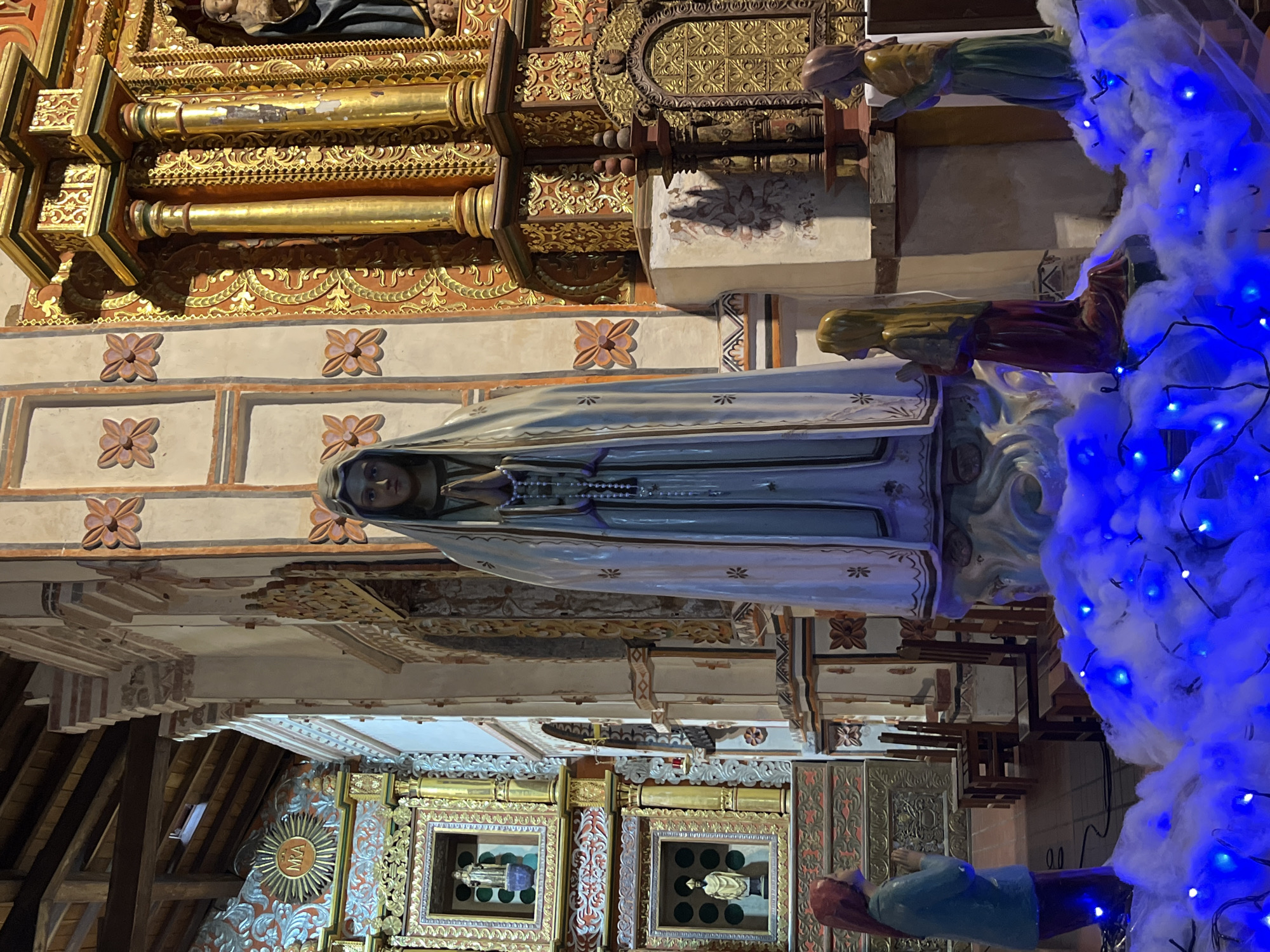 This image shows a statue of the Virgin Mary in what appears to be a church or religious setting. The statue is draped in a light-colored cloak and holding a rosary. It is surrounded by decorations including what seems to be cotton and blue LED lights. The architecture surrounding the statue includes ornate gold detailing and carvings, with repeated flower motifs on the walls. In the lower part of the image, there are smaller framed artworks on the wall. The precise location is unknown.