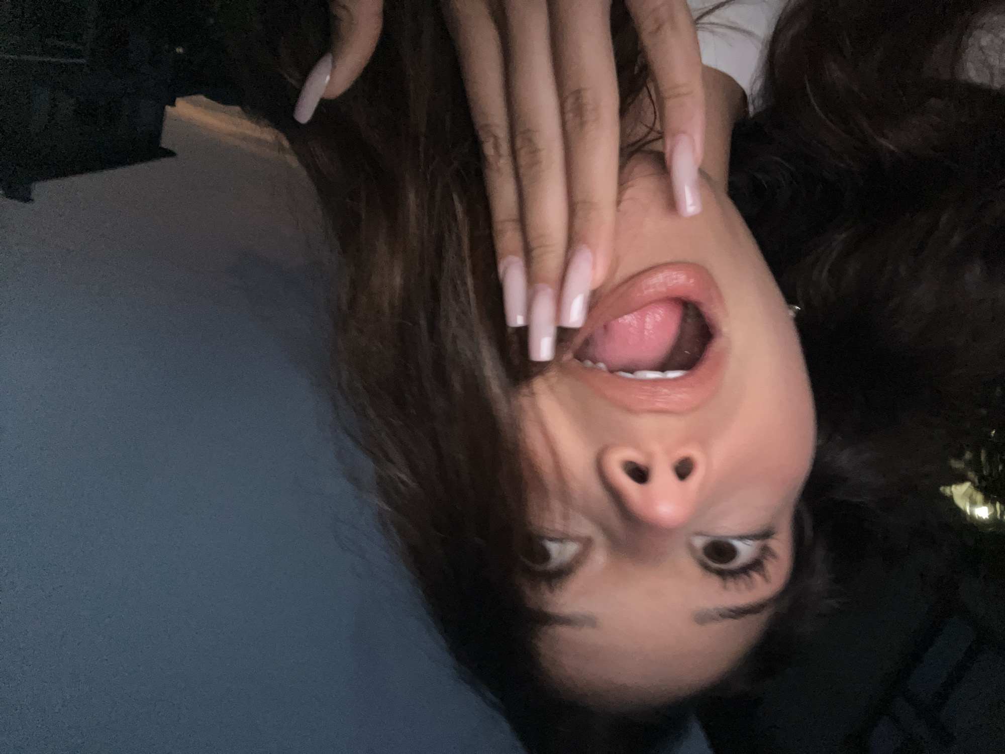 This media file is an upside-down, close-up, indoor selfie of a young woman with long, dark brown hair. She is wearing a white top. Her left hand, with long, light pink acrylic nails, is covering the lower half of her face, except for her mouth and nose. Her mouth is open, revealing her tongue, and her lips are glossy. Her eyes are wide, and she appears to be looking to her right. The background is dark and out of focus, with a hint of what might be a light source on the right side, suggesting it might be evening or night. The image has a soft focus and the lighting is dim. There is no visible text. The woman's expression could be interpreted as playful or surprised. The blue area to the left of the woman might be clothing or a piece of furniture. Based on the provided location context, this image was taken in Zlatograd, Bulgaria.