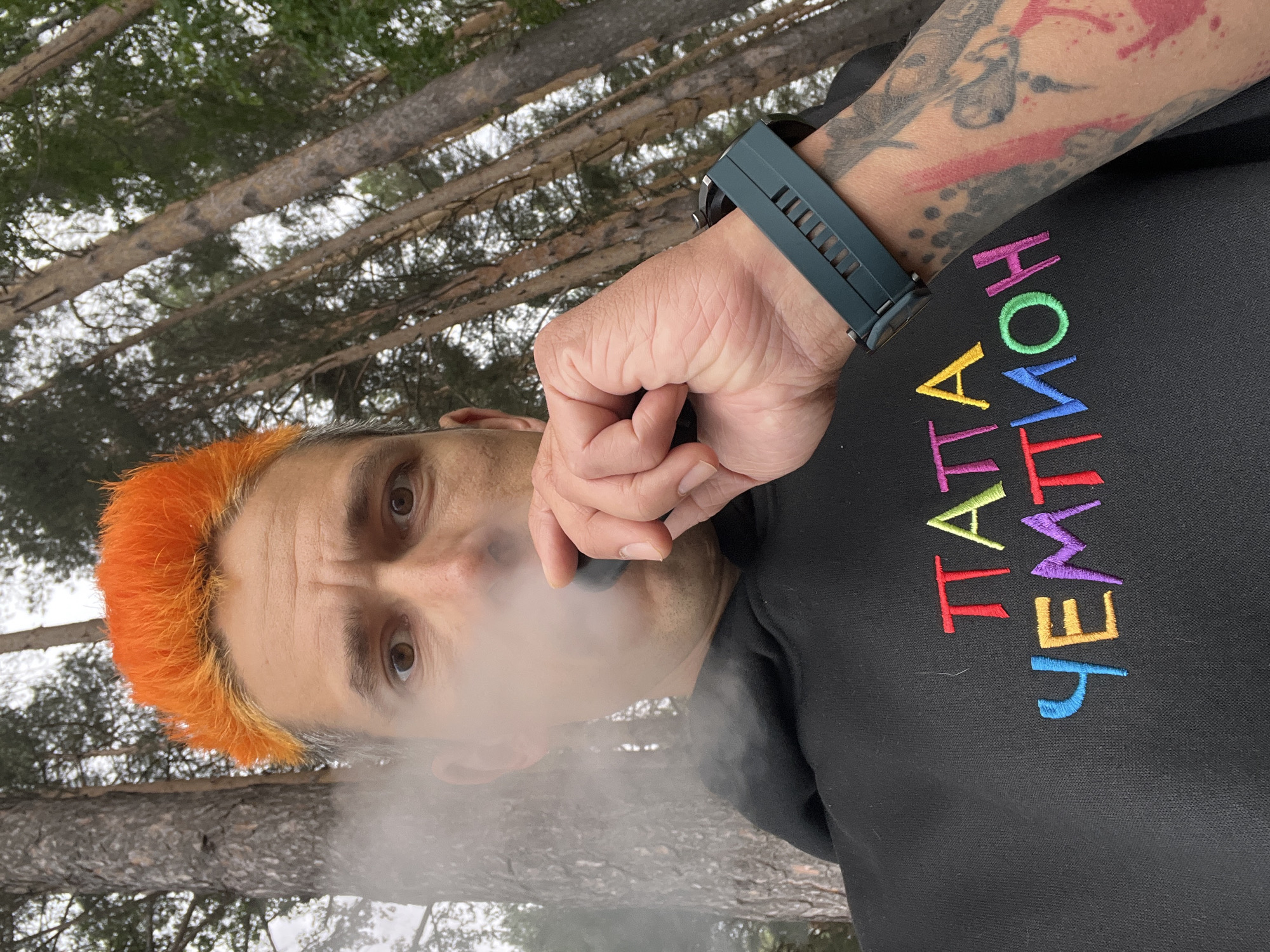 The image captures an adult male in a forested outdoor setting within the city of Chaikovsky, Russia. The man is depicted from a low-angle, medium close-up, looking directly into the camera while exhaling a cloud of white vapor from a dark, cylindrical vaping device held in his right hand. His hair is styled with a prominent bright orange, spiky top, transitioning to shorter, darker sides. He has brown eyes and a subtly furrowed brow. He wears a black hooded sweatshirt (hoodie) featuring the Cyrillic text "ПАПА ЧЕМПИОН" (Papa Champion) embroidered across the chest in multi-colored thread: 'П' in red, 'А' in orange, 'П' in purple, 'А' in greenish-yellow, 'Ч' in light blue, 'Е' in yellow, 'М' in purple, 'П' in pink, 'И' in blue, 'О' in green, and 'Н' in pink. A dark teal-colored smartwatch with a ribbed silicone band is visible on his left wrist, and his left forearm displays extensive tattoos featuring black outlines, grey shading, and splashes of red/pink. The background consists of numerous tall, slender conifer trees with textured bark, indicative of a woodland environment, under diffuse natural lighting suggesting an overcast sky.