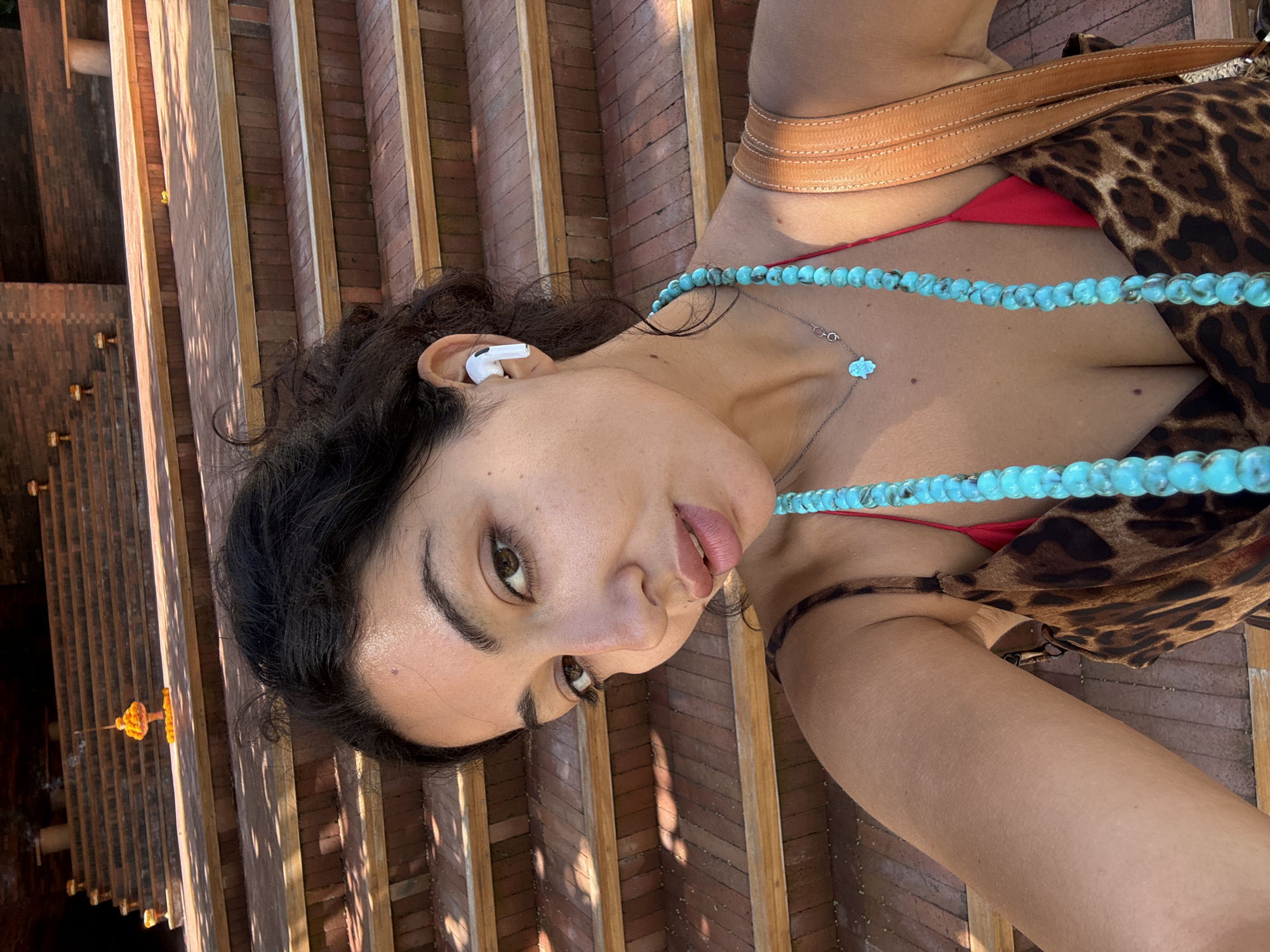 A woman wearing a red bikini top, a leopard-print coverup, and a turquoise necklace is lying on a wooden structure. She has dark hair and is wearing a white wireless earbud in her left ear. The structure appears to be a series of wooden planks with shadows cast across them, suggesting it is outdoors in bright sunlight. Based on the prompt information, the location is North Kuta, Indonesia.