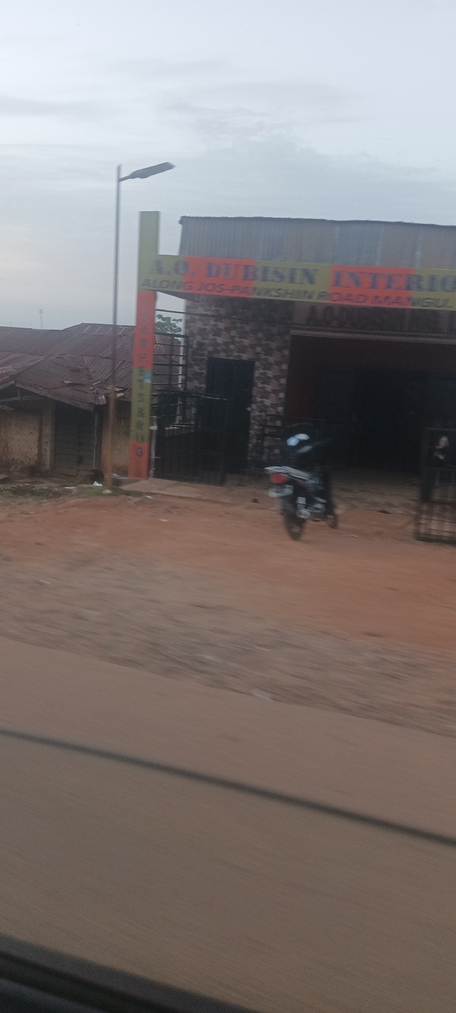 The image depicts a street view in Mangu Town, Nigeria. A building with a sign reading "A.O. DUBISIN INTERIOR ALONG JOS-PANKSHIN ROAD MANGU" stands alongside the road. The building appears to be a shop or business, possibly dealing in interior design or related services. A motorcycle is parked in front of the building. A separate, smaller structure is partially visible on the left side of the image. A street lamp is visible. The street is unpaved. The sky is overcast.
