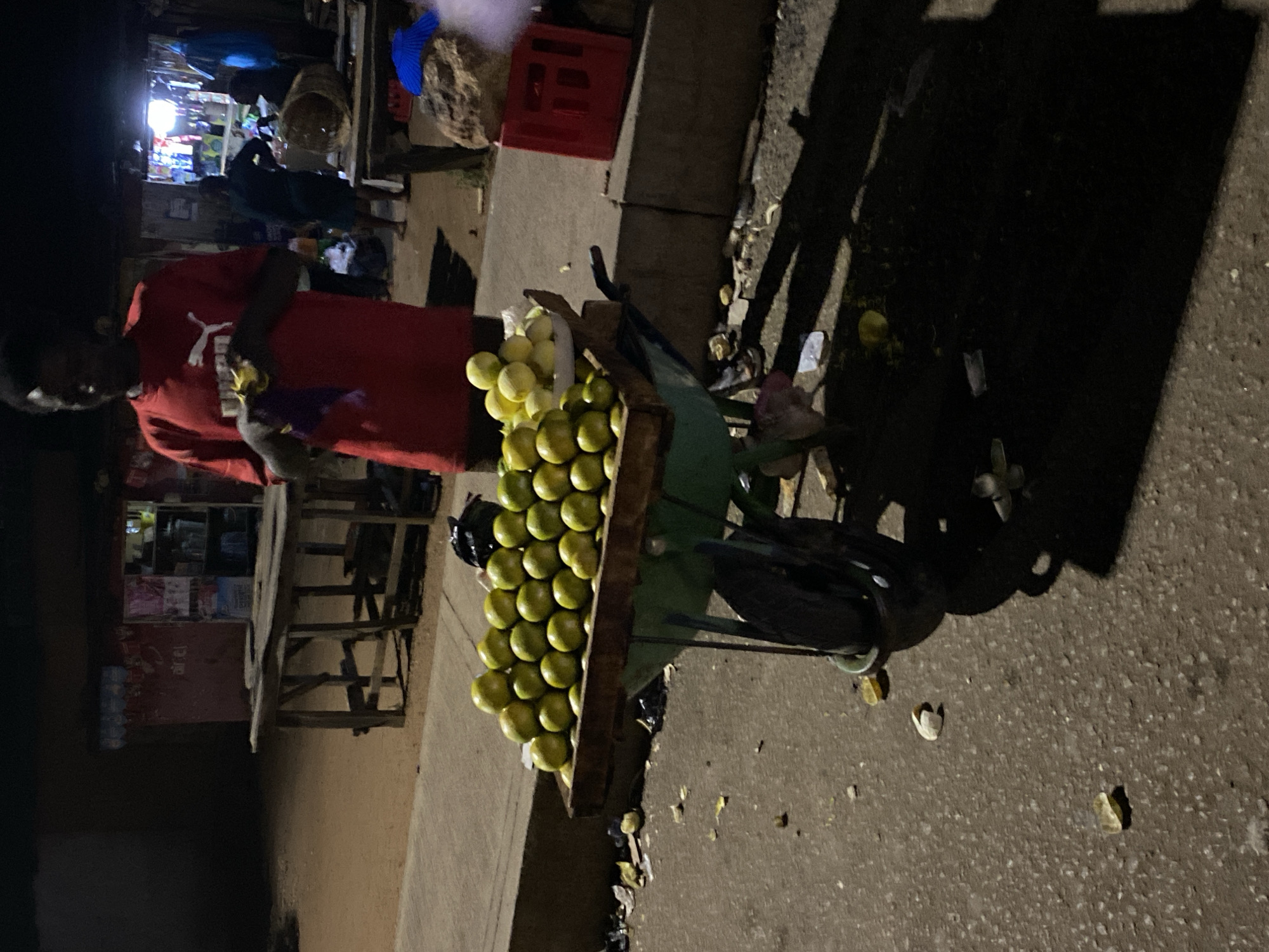 A street vendor stands next to a cart piled high with citrus fruit on a darkened street in Bukuru, Nigeria. The vendor, wearing a red shirt with a white Puma logo, appears to be interacting with a customer. The vendor's cart, a modified bicycle, is overloaded with pale green/yellow fruit. The scene is lit by dim ambient light, casting long shadows. A small shop with lighted shelves is visible in the background, next to a wooden table and chairs. There is discarded fruit waste on the ground near the cart. A red plastic crate is visible behind the cart.