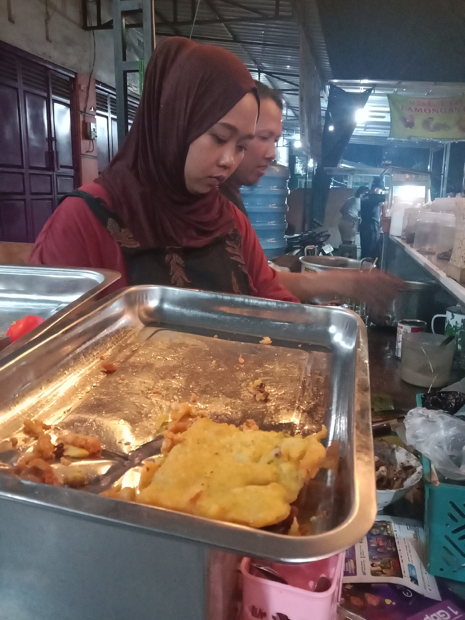 In Cangkring, Indonesia, a food stall vendor, a woman wearing a maroon hijab and a red shirt with a dark apron, is preparing food. She is standing behind a stainless steel serving tray filled with fried items and is using tongs. To her left is another stainless steel tray. Behind her, a man with short hair, presumably a colleague or customer, is visible. The stall appears to be outdoors, under a corrugated metal roof, with a sign in the background that reads "LAMONGAN". Various containers, a large water cooler, and bags are present in the background, suggesting a busy food service environment.