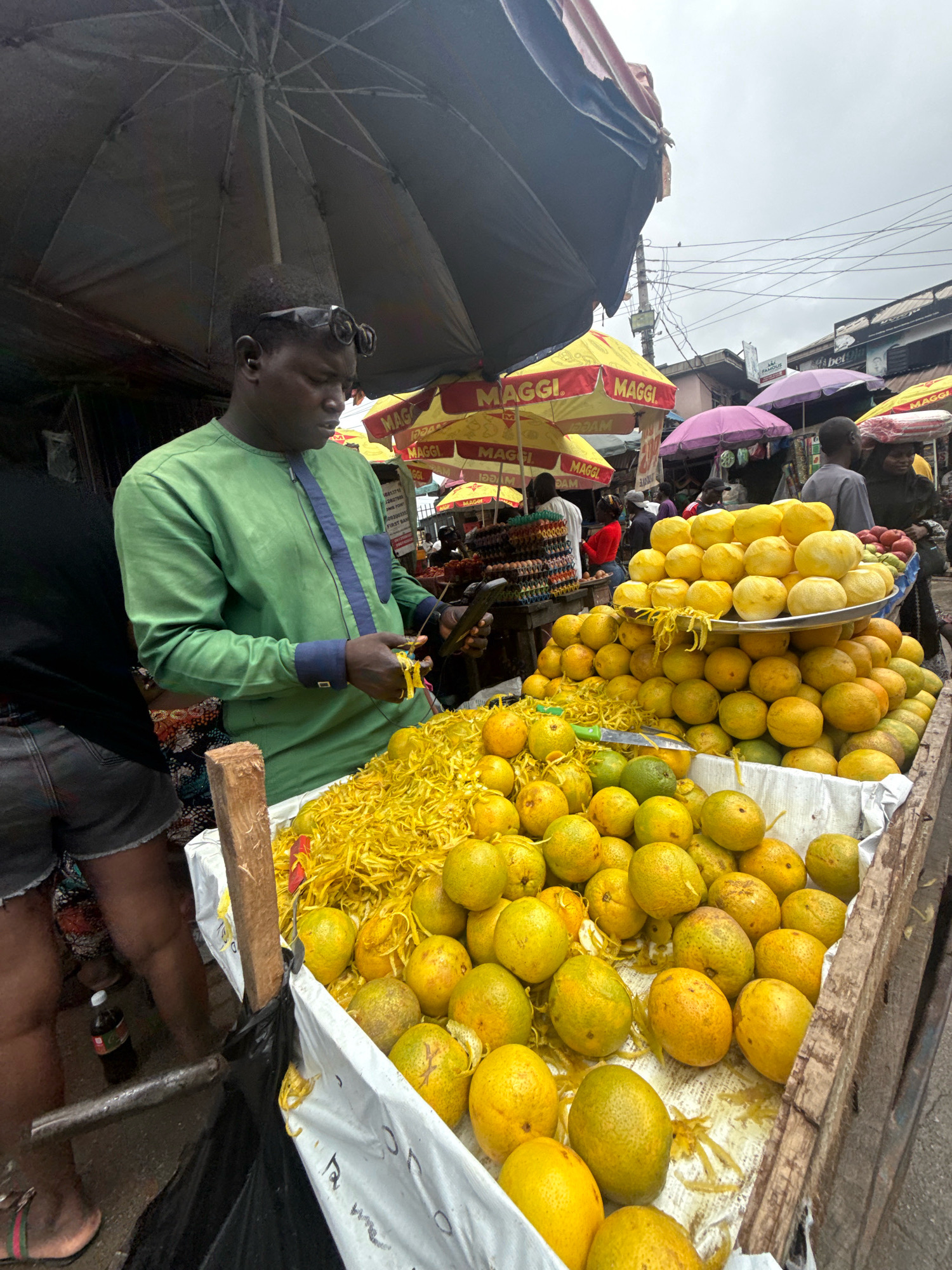 In Ado, Nigeria, a man in a green tunic with blue trim is preparing what appears to be shredded citrus peels at a market stall overflowing with oranges. He holds a knife and a bunch of peels in his hands, actively engaged in his task. The stall is laden with a large quantity of fresh oranges, some piled on a metal tray and many more spilling out onto the cart. Scattered amongst the oranges are heaps of the bright yellow, shredded peel.
Behind the fruit seller, various market stalls are visible, adorned with umbrellas, some featuring the "MAGGI" logo. Other vendors and shoppers can be seen in the background, suggesting a bustling marketplace environment. The sky is overcast, hinting at the possibility of rain or a generally cloudy day. The scene depicts a typical street market in Ado, highlighting local commerce and the availability of fresh produce.