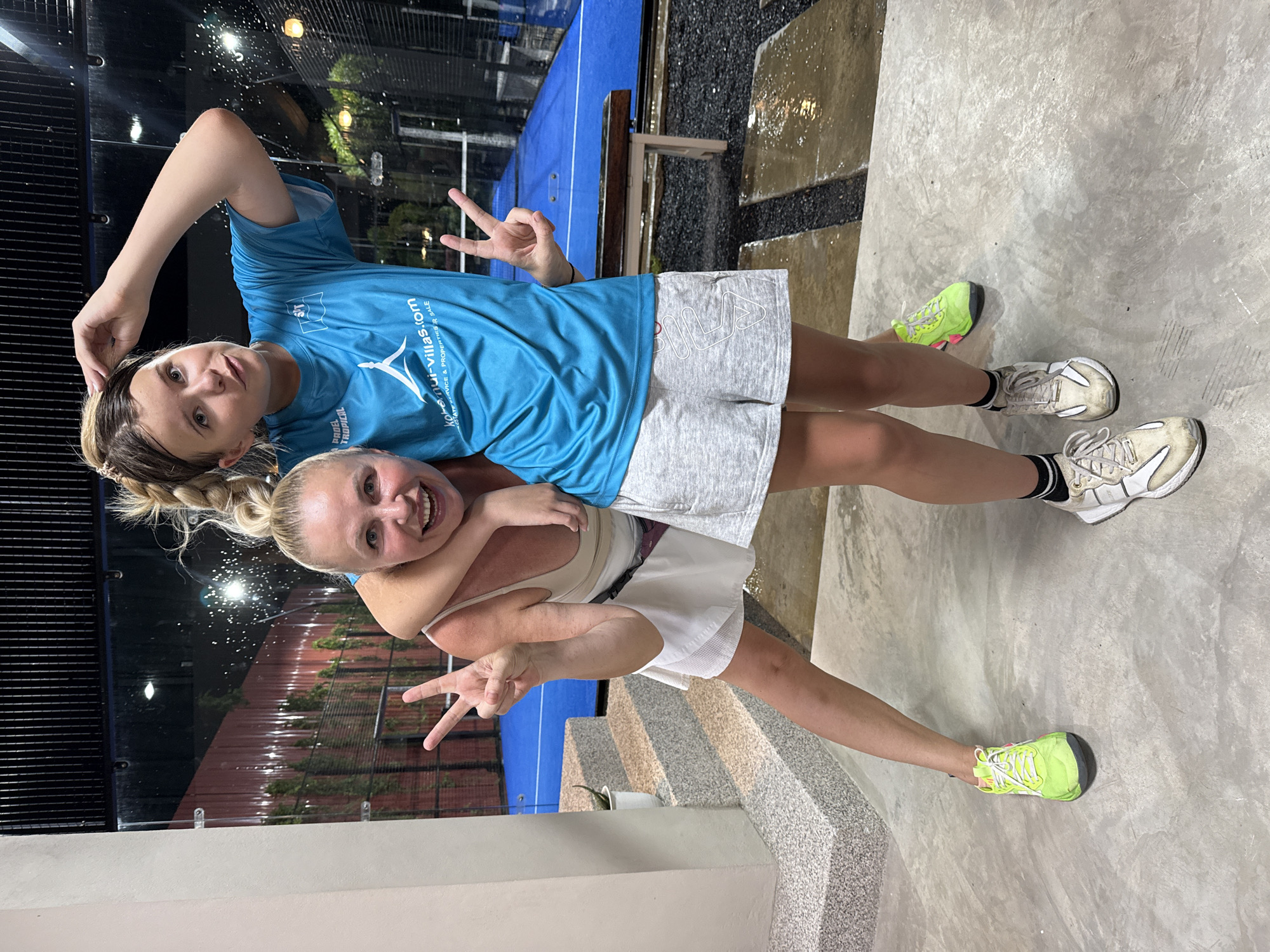 Two women pose for a photo on a concrete floor with a blue padel court in the background. The woman in the foreground, on the left, has blonde hair pulled back in a ponytail and is wearing a light-colored tank top, a white skirt, and lime green athletic shoes. She is embracing the woman behind her from the front and is making a peace sign with her left hand. The woman behind her, on the right, has blonde hair in a braid and is wearing a bright blue t-shirt and gray athletic shorts with white athletic shoes. She has her right arm around the neck of the woman in front of her and is touching her head with her left hand while making a peace sign with her right hand. The background shows a dimly lit padel court with a blue surface and black netting, and a wooden bench. The environment appears to be an outdoor or semi-outdoor recreational facility, possibly a sports club or resort, located in Ko Samui District, Thailand, as indicated by the text on the t-shirt which reads "kohl-samui-villas.com". The ground is wet in places, suggesting recent rain or cleaning.