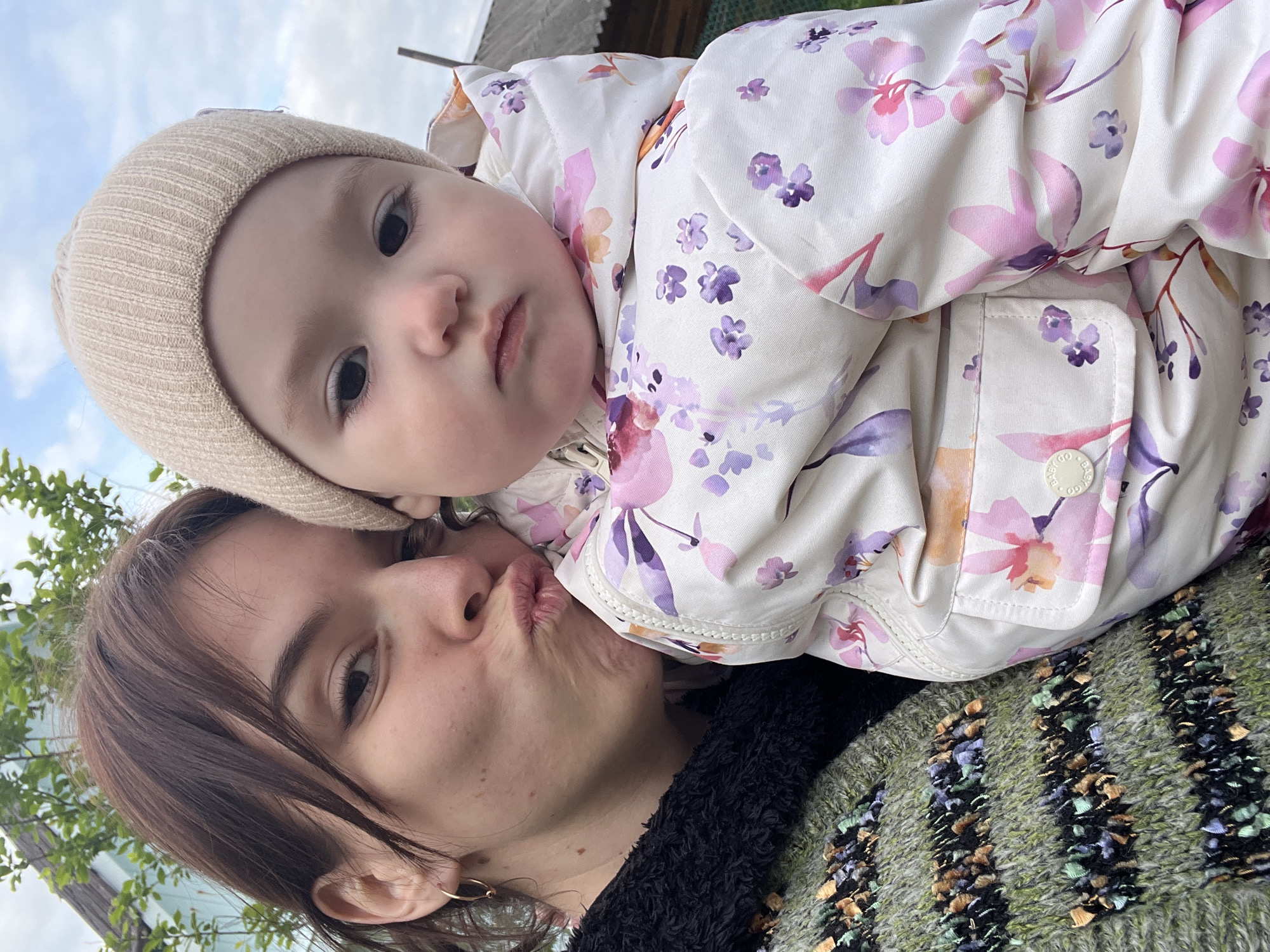 A woman and a baby are in a selfie taken outdoors, likely in Мшинская, Russia. The woman, with brown hair pulled back and a small gold hoop earring visible, is making a kissing face toward the camera. She is wearing a green and black textured sweater. The baby, wearing a beige knit hat and a white jacket with a purple and pink floral pattern, is resting on the woman. The background includes green foliage and a glimpse of a building. The sky is visible in the upper left corner.