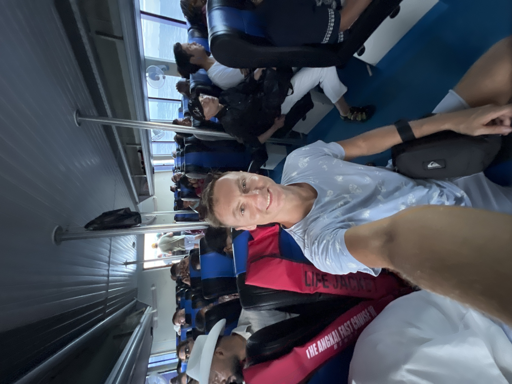 The image displays the interior of a fast cruise boat or ferry, captured from a low angle as a selfie. A smiling man in a light blue patterned t-shirt is in the foreground, holding the camera. A black fanny pack is visible on his lap or by his side.
Rows of blue upholstered seats line the cabin, occupied by numerous passengers. Many people are visible throughout the boat, some looking forward, others less distinct. Next to the smiling man, another passenger wears a white brimmed hat and a red life jacket. Visible text on the life jacket clearly reads "LIFE JACKET" and "THE ANGKAI FAST CRUISE 16," indicating the name of the ferry operator and potentially the boat number.
The boat's interior features a ribbed, paneled ceiling and multiple metal support poles. Natural light illuminates the cabin, suggesting it is daytime, filtering in through large windows that run along the side of the vessel. A small white fan is mounted near one of these windows. Through the windows, glimpses of the outside environment or another deck are visible. The atmosphere appears casual, with passengers undertaking a journey. The image is rotated 90 degrees clockwise. Given the location context of Denpasar, Indonesia, this scene likely depicts travel between islands.