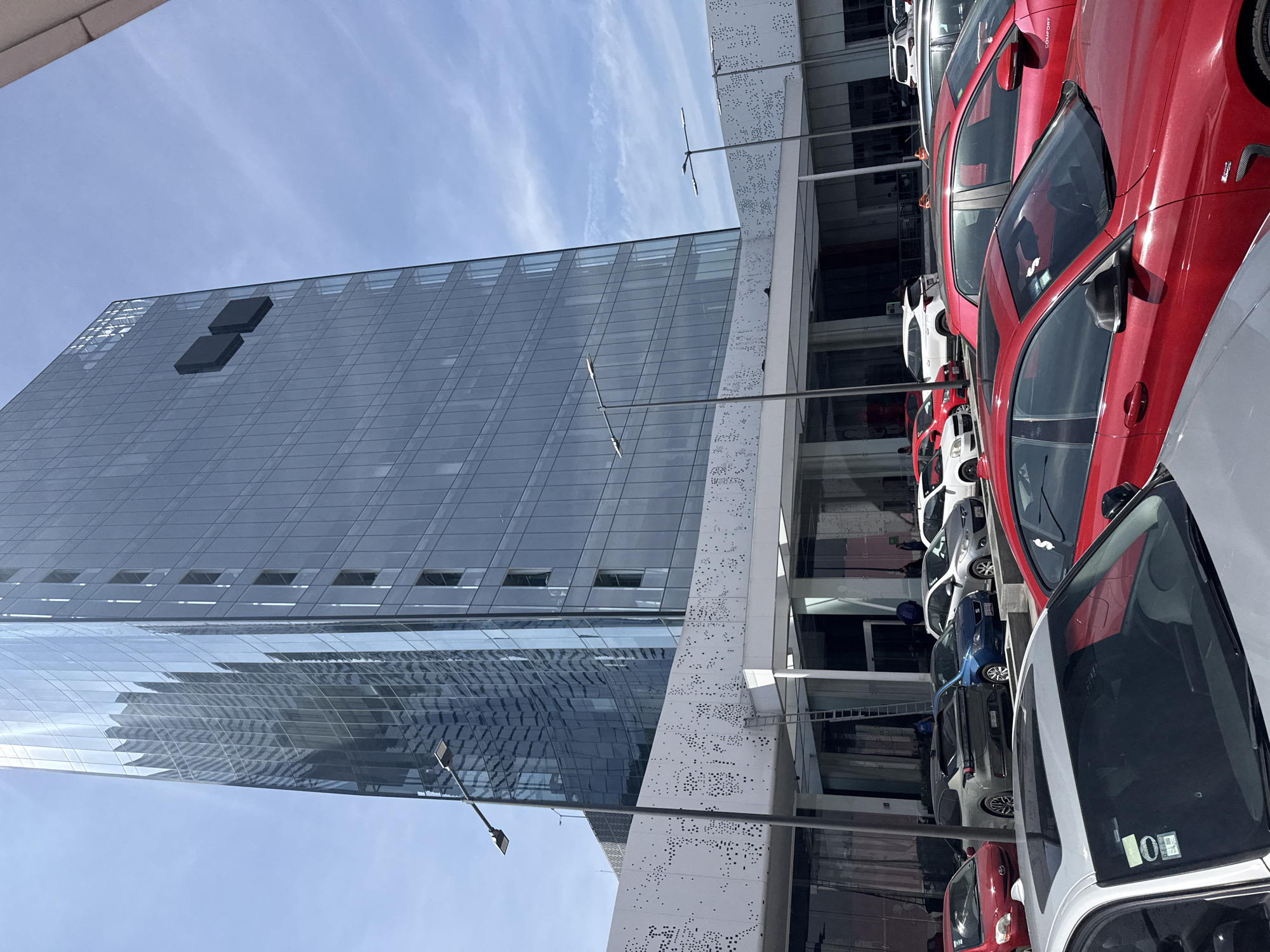 The image depicts a modern, glass-clad building in Puebla, Mexico, likely an office tower or commercial structure, with a secondary building alongside featuring a white facade with perforated designs. A row of parked cars in various colors (red, white, blue, and gray) is visible in the foreground. The presence of the cars suggests the location might be a parking area for the building or a nearby establishment. The sky is blue with sparse clouds, indicating fair weather. The image provides no information about people or interactions.