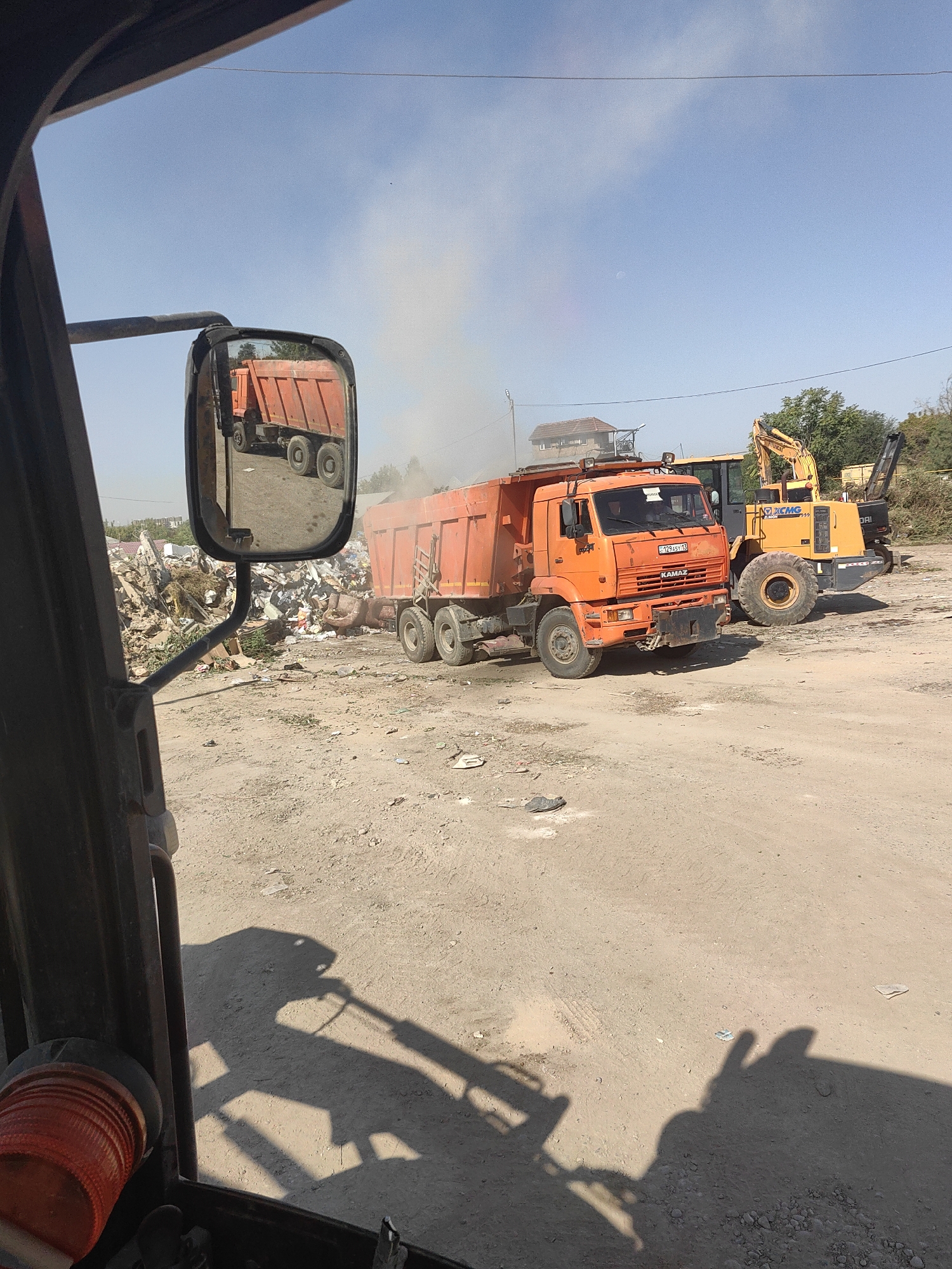 In Shymkent, Kazakhstan, an orange KAMAZ dump truck is parked on a dusty, unpaved lot filled with debris and construction waste. A yellow XCMG excavator is also present, positioned behind the dump truck. The dump truck's bed appears to be tilted upwards, and smoke or dust is rising from it, suggesting recent activity. A side mirror of a vehicle, likely the one from which the photo is taken, reflects the dump truck and the surrounding junk pile. The sky is clear and blue, with a few wispy clouds. The environment appears to be an open-air waste disposal or recycling area.