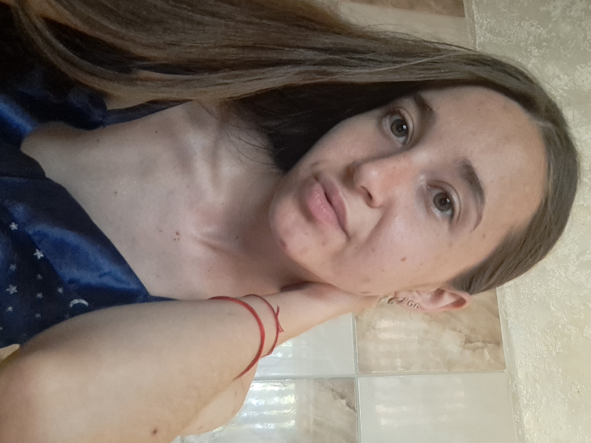 A young woman with long brown hair is captured in a close-up, selfie-style shot. She is wearing a blue silk top with a celestial pattern. A thin red string bracelet is visible on her left wrist. Her left arm is bent and her hand is touching the back of her neck, while her head is tilted slightly to her right. The background appears to be a tiled wall, possibly in a bathroom. The image does not provide any information about the city of Vypasne, Ukraine, or any other geographical details.