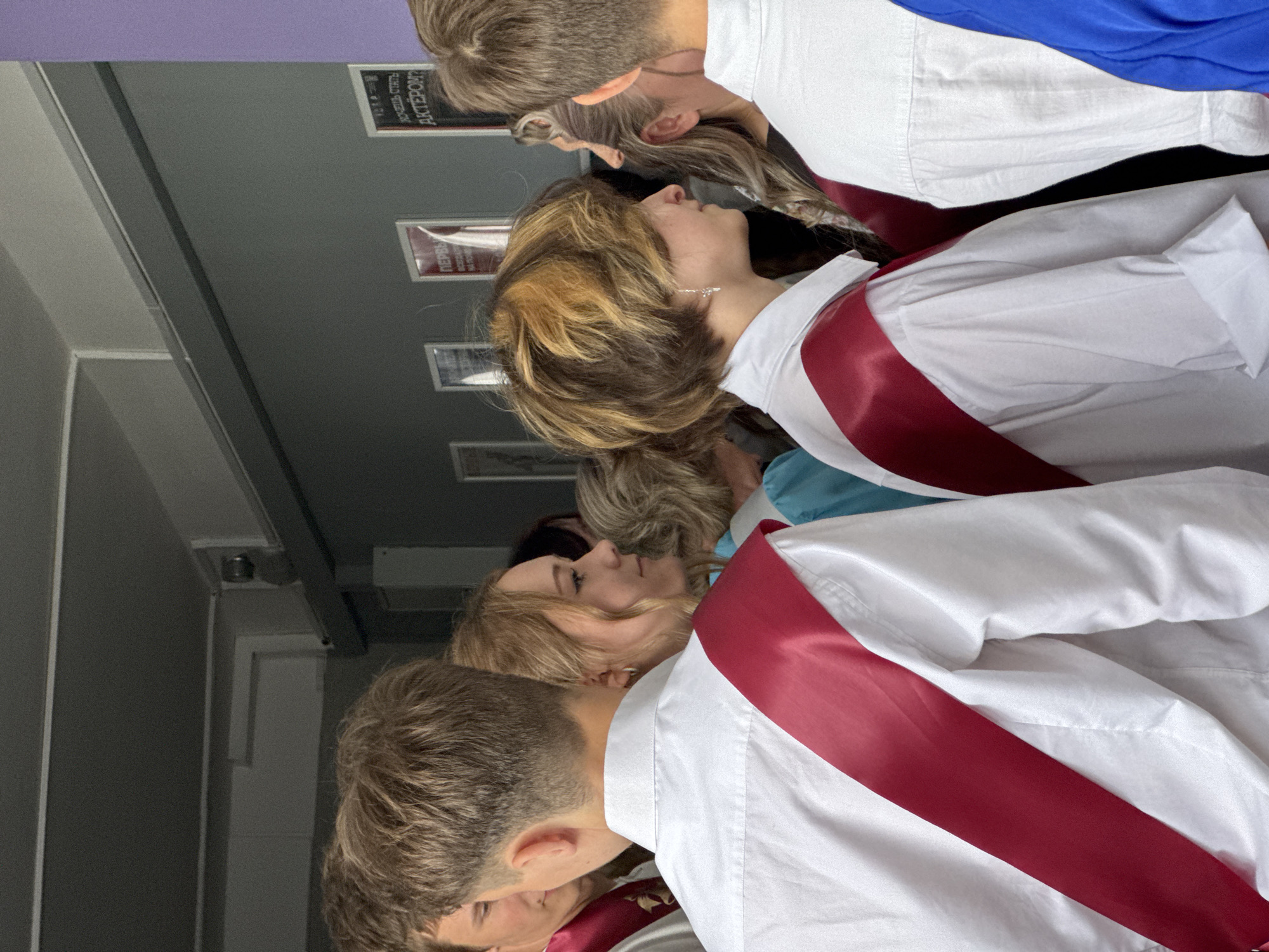 The image shows a group of students, likely graduates, standing indoors in a hallway. They are wearing white shirts with red sashes. The location appears to be Прикамский, Russia, based on the signs visible in the background.