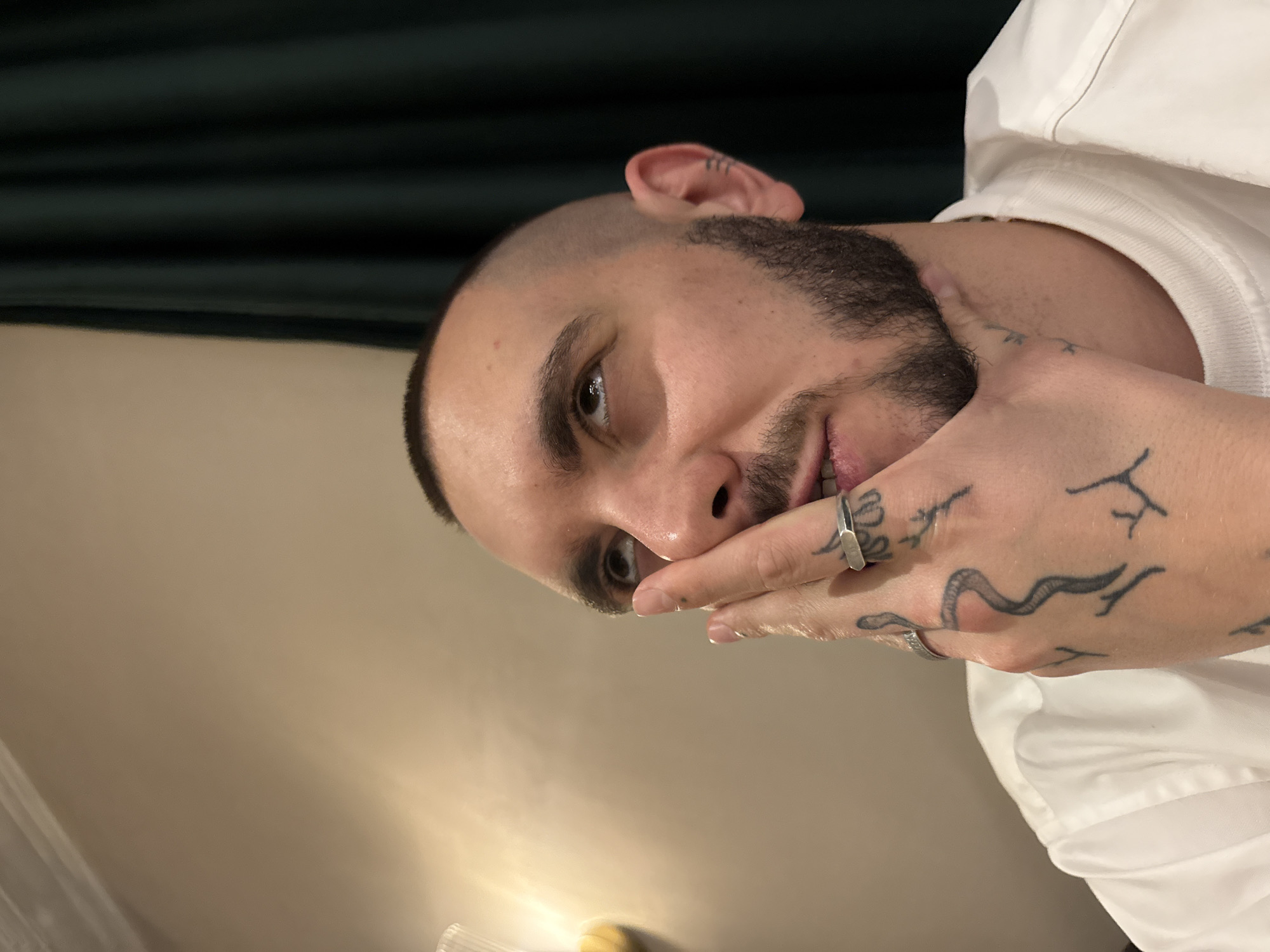A close-up, rotated image shows a man with short, dark hair and a beard. His hand is raised to his face, with fingers partially covering his mouth. He has tattoos on his hand and wears two rings. The background includes a dark green surface and a light-colored wall. The location in Yerevan, Armenia is not discernible from the image.