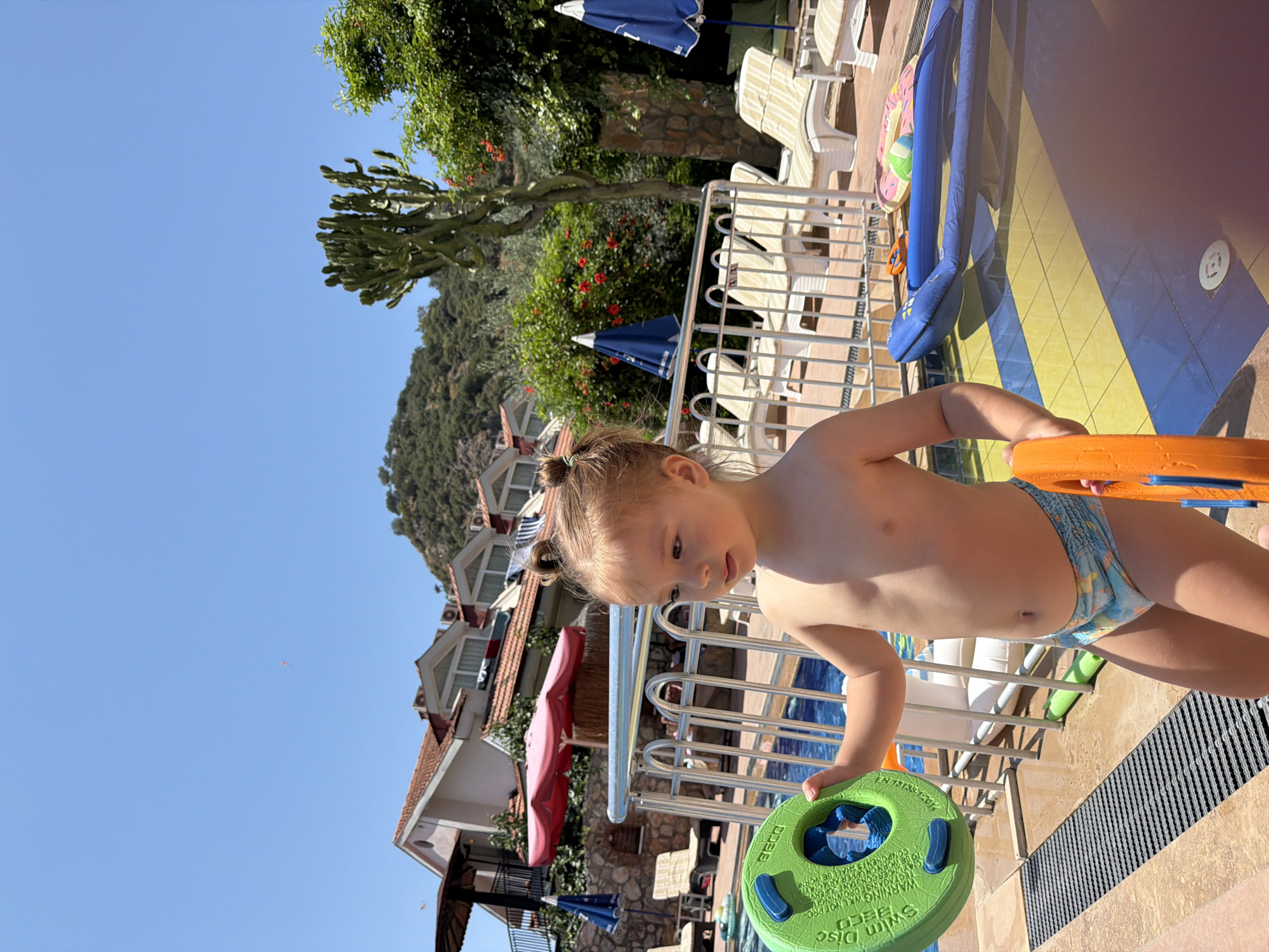 A young child stands near a swimming pool, holding a green flotation device. The child is wearing swimming trunks and has their hair in two small buns. In the background, there are lounge chairs, umbrellas, and buildings that appear to be part of a resort. The scene is set outdoors under a clear blue sky, suggesting a sunny day. The buildings and lush greenery in the background are consistent with the landscape of Fethiye, Turkey.