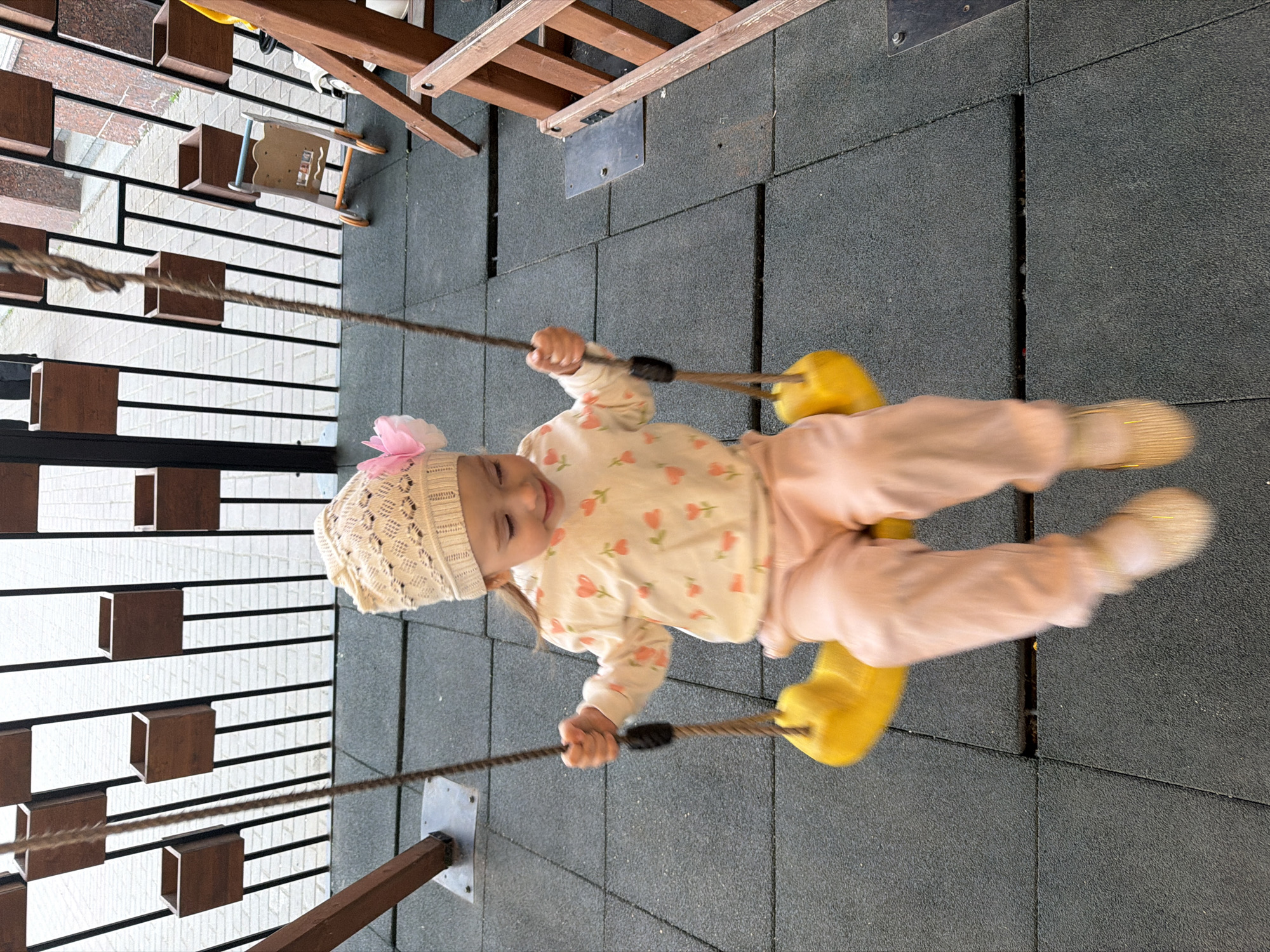 In this medium shot, a young child is captured mid-swing on a playground. The child, wearing a light-colored knit hat adorned with a pink flower and a long-sleeved shirt with a heart pattern, is swinging on a swing with bright yellow handles. They are wearing light pink pants and yellow shoes. The child's eyes are closed, and their face conveys a sense of joy and exhilaration as they are propelled through the air.
The playground features a dark grey rubberized surface, typical of modern play areas designed for safety. In the background, a wooden structure, likely part of the swing set or another playground apparatus, is visible. To the left, a fence-like structure with horizontal wooden slats and brown rectangular boxes adds to the visual context of the outdoor setting. The lighting suggests it is daytime, and the scene appears to be set in a park or a dedicated children's play area. The overall atmosphere is one of cheerful activity and childhood enjoyment.