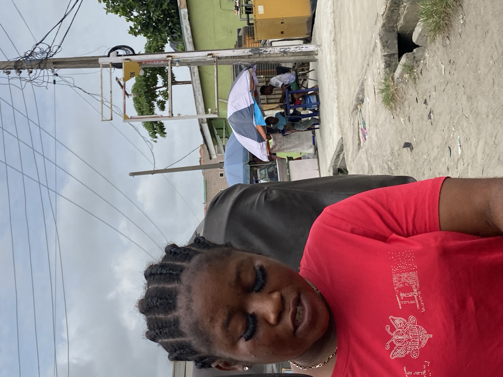 The image shows a woman with braided hair and a red t-shirt. In the background are figures under umbrellas, seated outdoors. Also in the scene is a concrete surface. The location appears to be outdoors, possibly in Ado, Nigeria.