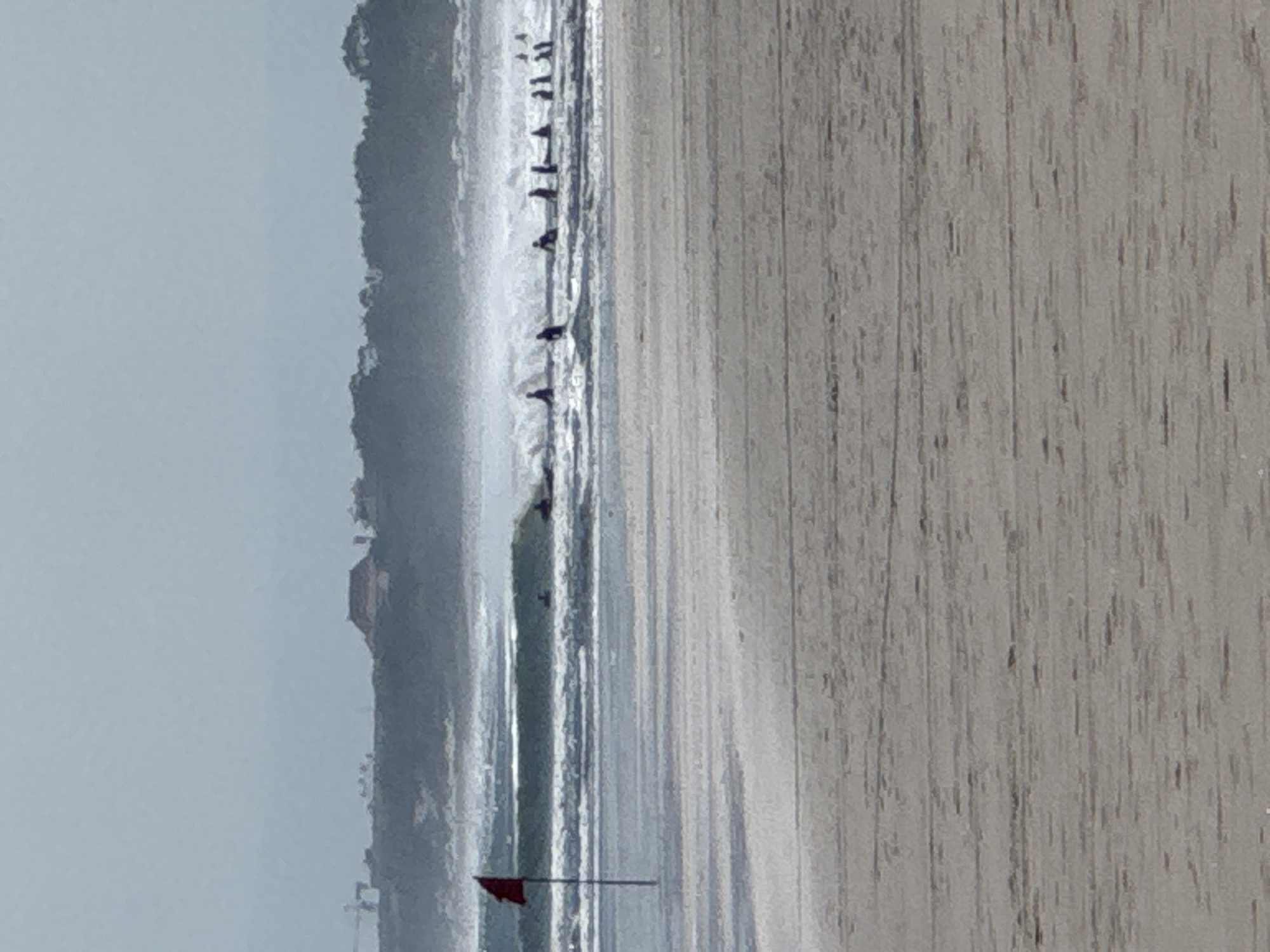A wide shot of Kuta Beach, Bali, Indonesia captures a hazy, overcast day. In the foreground, the sandy beach stretches towards the ocean. A red flag on a pole stands prominently on the left, marking the edge of the surf. The ocean itself features moderate waves, with several surfers visible in the water, some riding waves and others waiting. In the distance, a tree-lined coast forms the backdrop, with a cluster of buildings, possibly resorts or homes, nestled amongst the trees. The overall atmosphere is serene, with the muted colors and soft focus contributing to a tranquil beach scene.