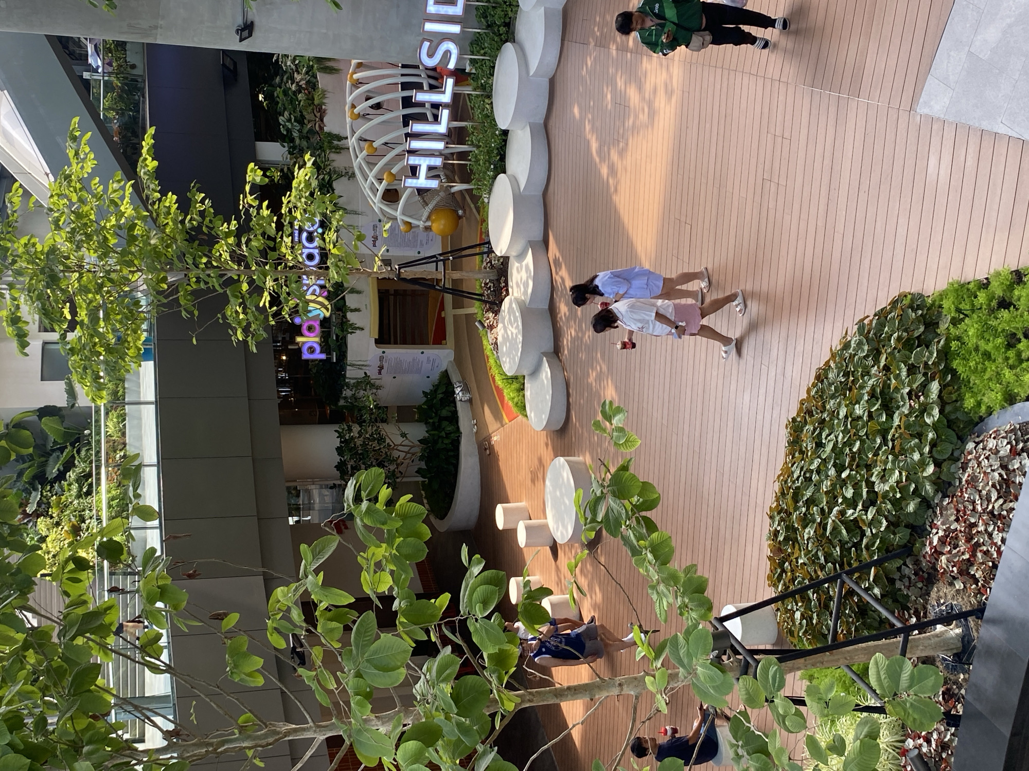 Here is a description of the image:
The image shows an outdoor courtyard area in Bangkok, Thailand. The word "HILLSIDE" is written in large white letters on a structure in the background. A sign that reads "playspace" in bright purple lettering is visible near a building. There are several people in the image; two girls are walking together, one person is wearing a green shirt, and several people are sitting and conversing in a seating area. The courtyard features wooden-look flooring, various plants and greenery, and circular white structures that appear to be seating. The image is taken from an elevated position, partially obscured by tree branches in the foreground.