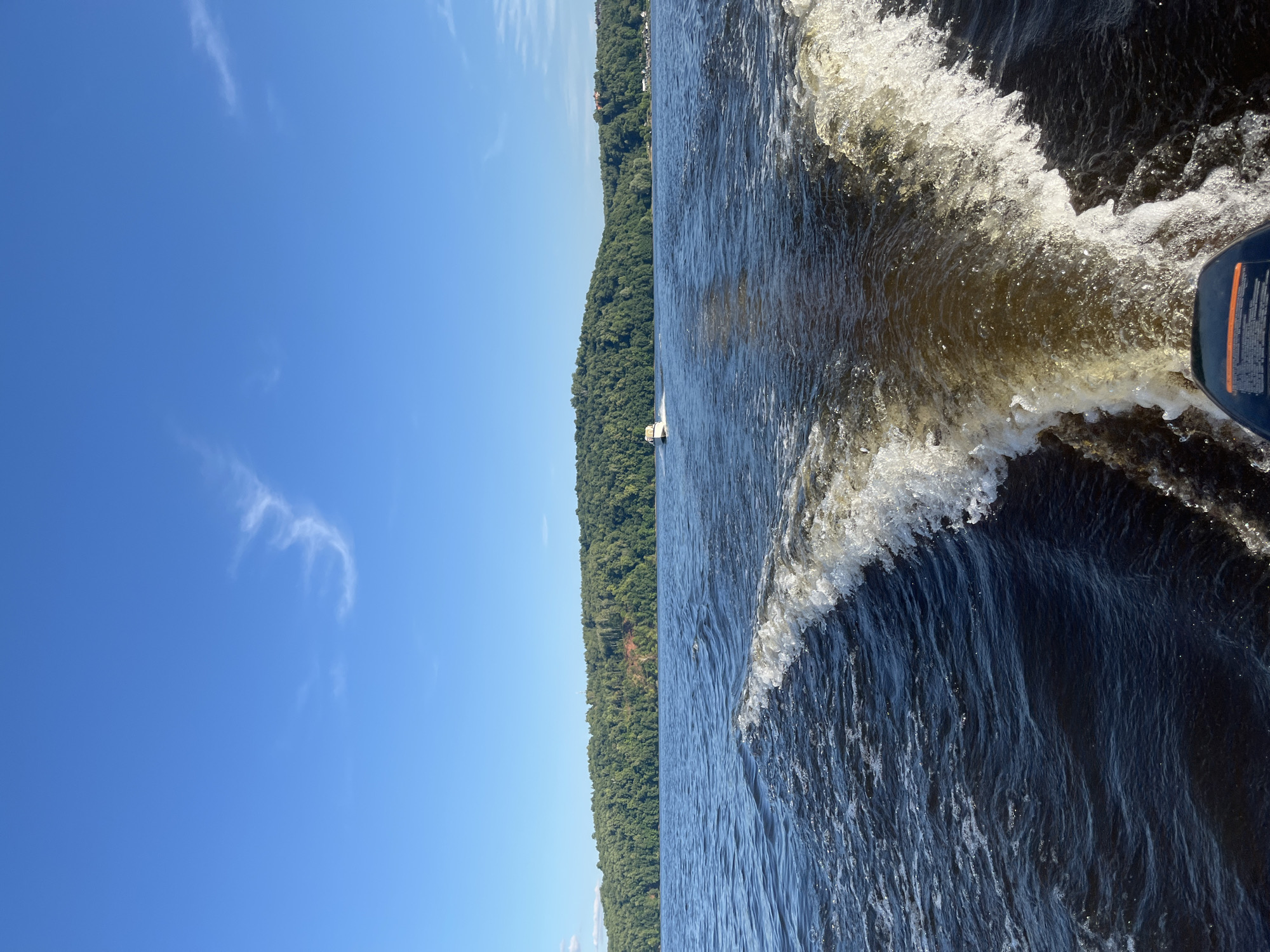 The image depicts a scenic view from a boat moving through a body of water, likely a river or lake, with a densely forested shoreline. The perspective is from the stern of the boat, looking towards the left and slightly forward. The water is dark blue and disturbed by the boat's wake, creating foamy white crests and ripples.
The shoreline is covered in a thick, green forest that extends to the horizon. A clear, bright blue sky with a few wispy white clouds dominates the upper left portion of the image. The sun appears to be high in the sky, suggesting it is daytime, likely mid-morning or afternoon, with good weather.
A small white boat is visible in the distance near the shoreline. The motor of the boat from which the photo is taken is partially visible in the lower right corner, showing a blue housing with some orange accents and text that is mostly illegible due to glare and focus.
The overall mood is peaceful and serene, capturing a moment of leisurely travel through a natural landscape. The location is identified as Siukterka, Russia.