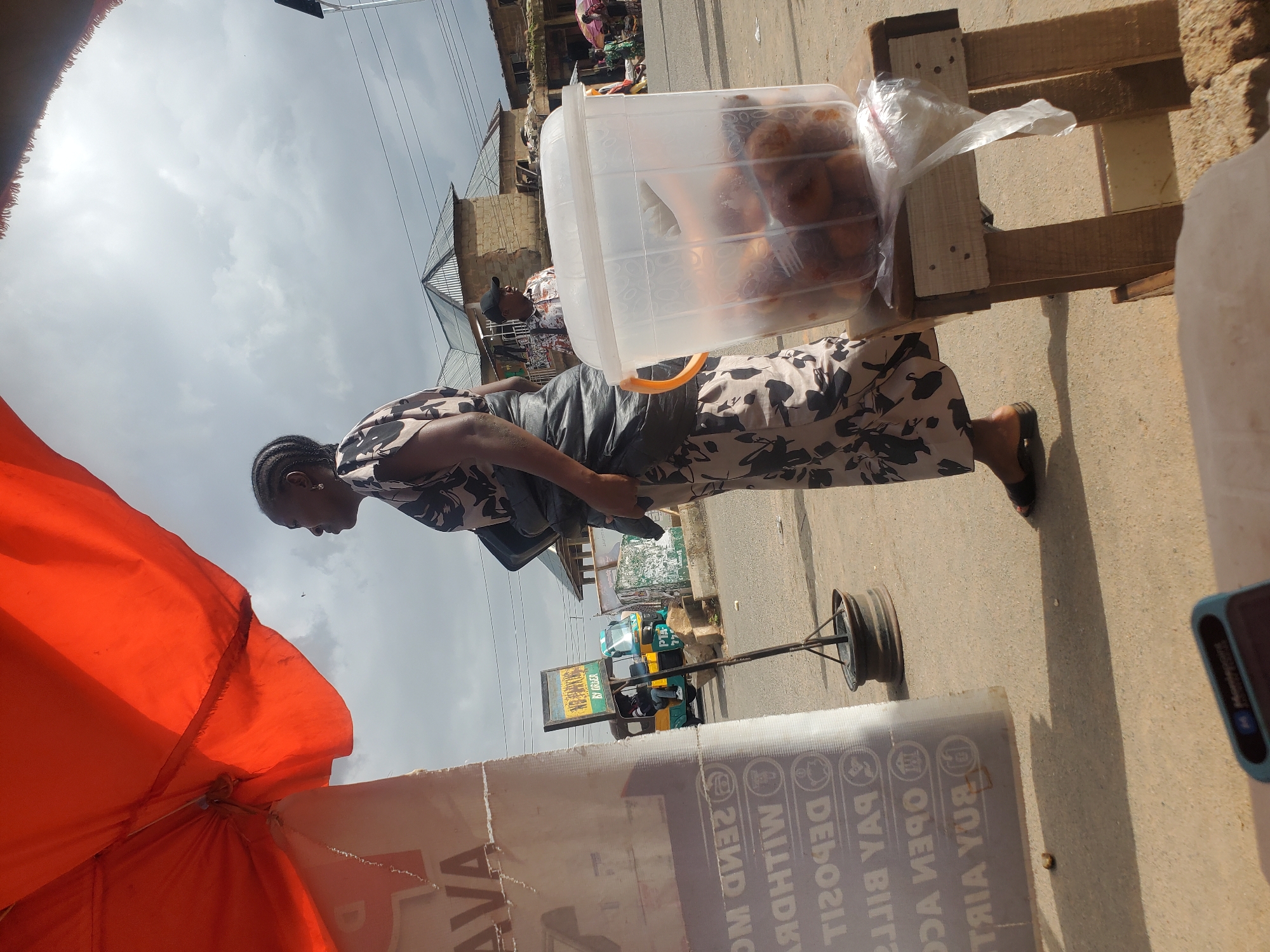 A woman stands next to a clear plastic container filled with fried snacks, likely doughnuts, displayed on a wooden stand. She is wearing a black and white patterned dress and dark sandals. Behind her, a man in a checkered shirt and a black cap is visible, along with other market stalls and buildings. An orange umbrella is partially visible in the foreground. A rickshaw, possibly a commercial vehicle, with an advertisement board, is in the background. The sky is overcast, suggesting a cloudy day. The scene appears to be a marketplace in Gwong, Nigeria. Visible text includes "SEND MONEY," "DEPOSIT," "PAY BILLS," "OPEN ACCOUNTS," and "BUY AIRTIME" on a sign partially obscured by the umbrella.
