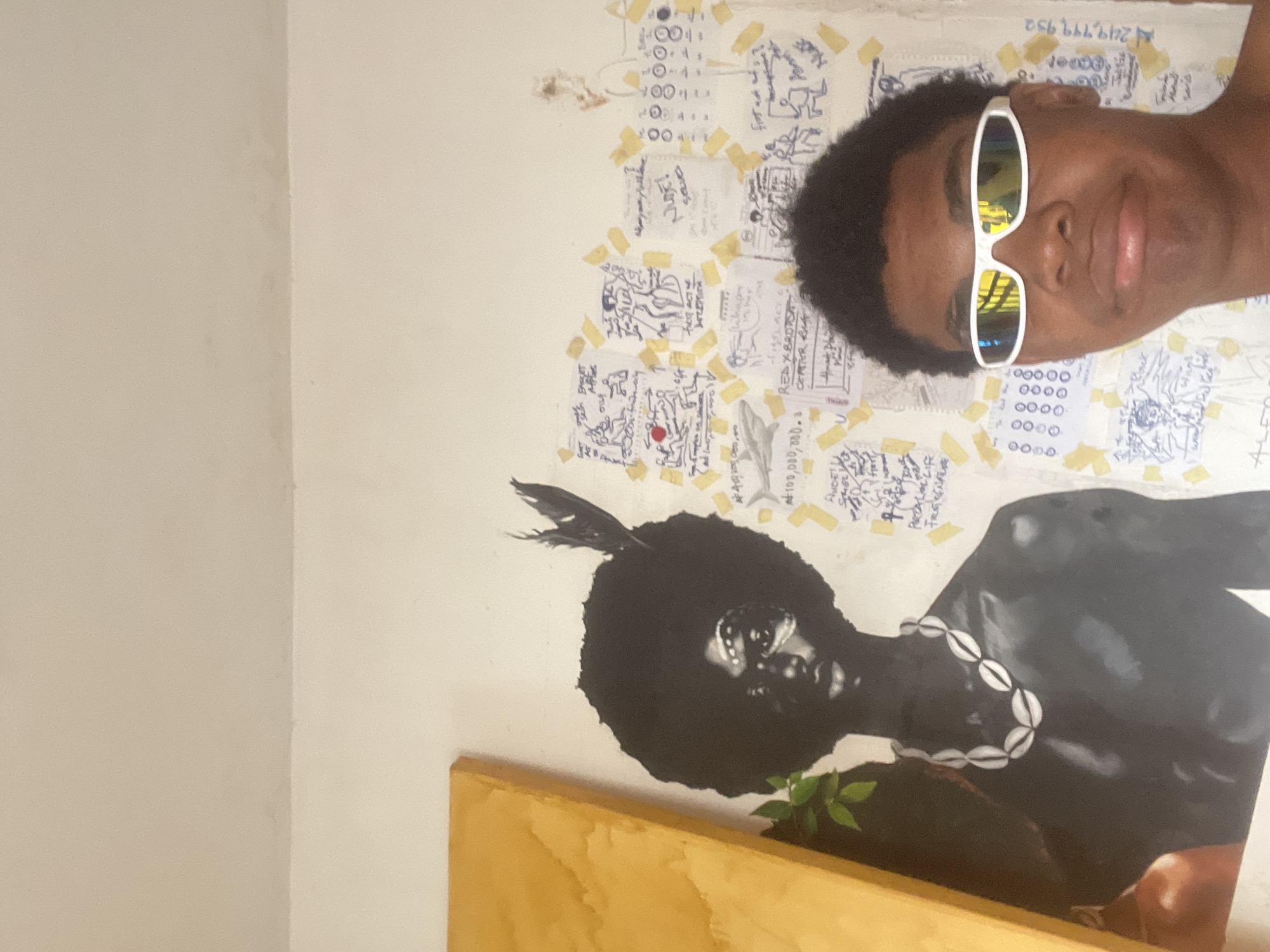 The image shows a young person wearing white-rimmed sunglasses, taking a selfie in front of a wall. Behind the person, there's a painting of a dark-skinned individual with an afro and a shell necklace, adorned with white markings on their face and a feather in their hair. The wall is covered with various papers, some with handwritten text and drawings, taped up with yellow tape. To the left of the painting, a portion of a large, yellow canvas is visible. The overall impression is of an indoor space, possibly an artist's studio or a personal living area, given the artwork and the eclectic arrangement of papers. The specific location in Ado, Nigeria, cannot be determined from the visual information provided.