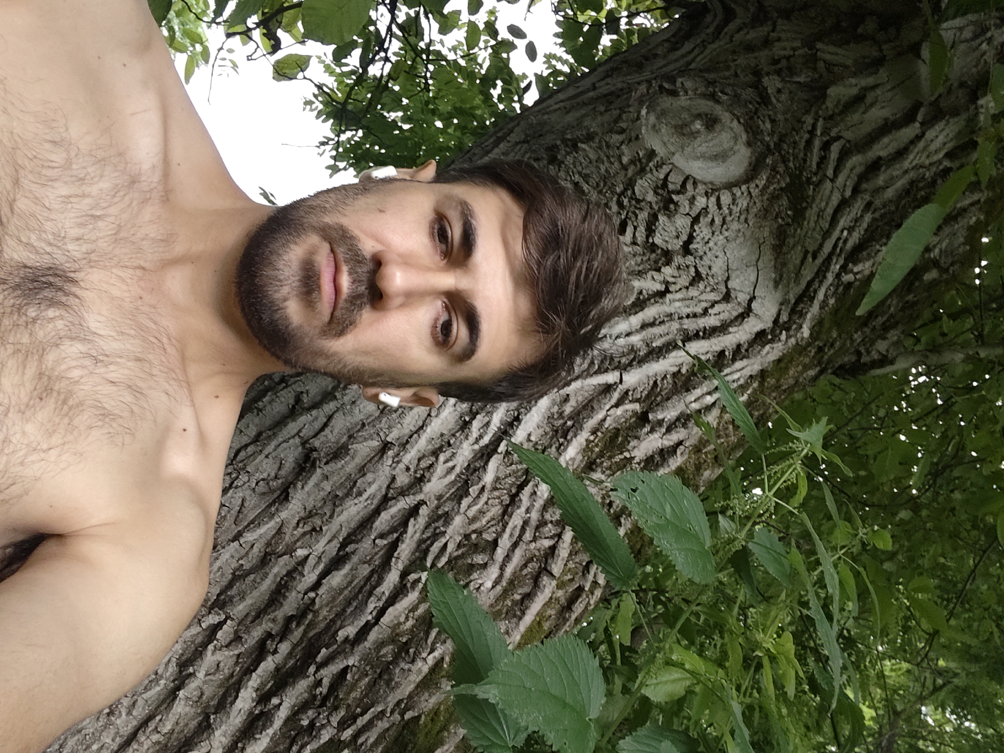 The image shows a close-up, low-angle shot of a man lying on his back against a large tree trunk. The man is shirtless, revealing a hairy chest and shoulder. He has a beard and is wearing a white wireless earbud in his left ear. His eyes are open and looking towards the camera. Green leaves and branches of trees are visible in the background, with a bright sky peeking through the foliage. The tree bark is rough and textured. The location is identified as Akhmetovskaya, Russia.