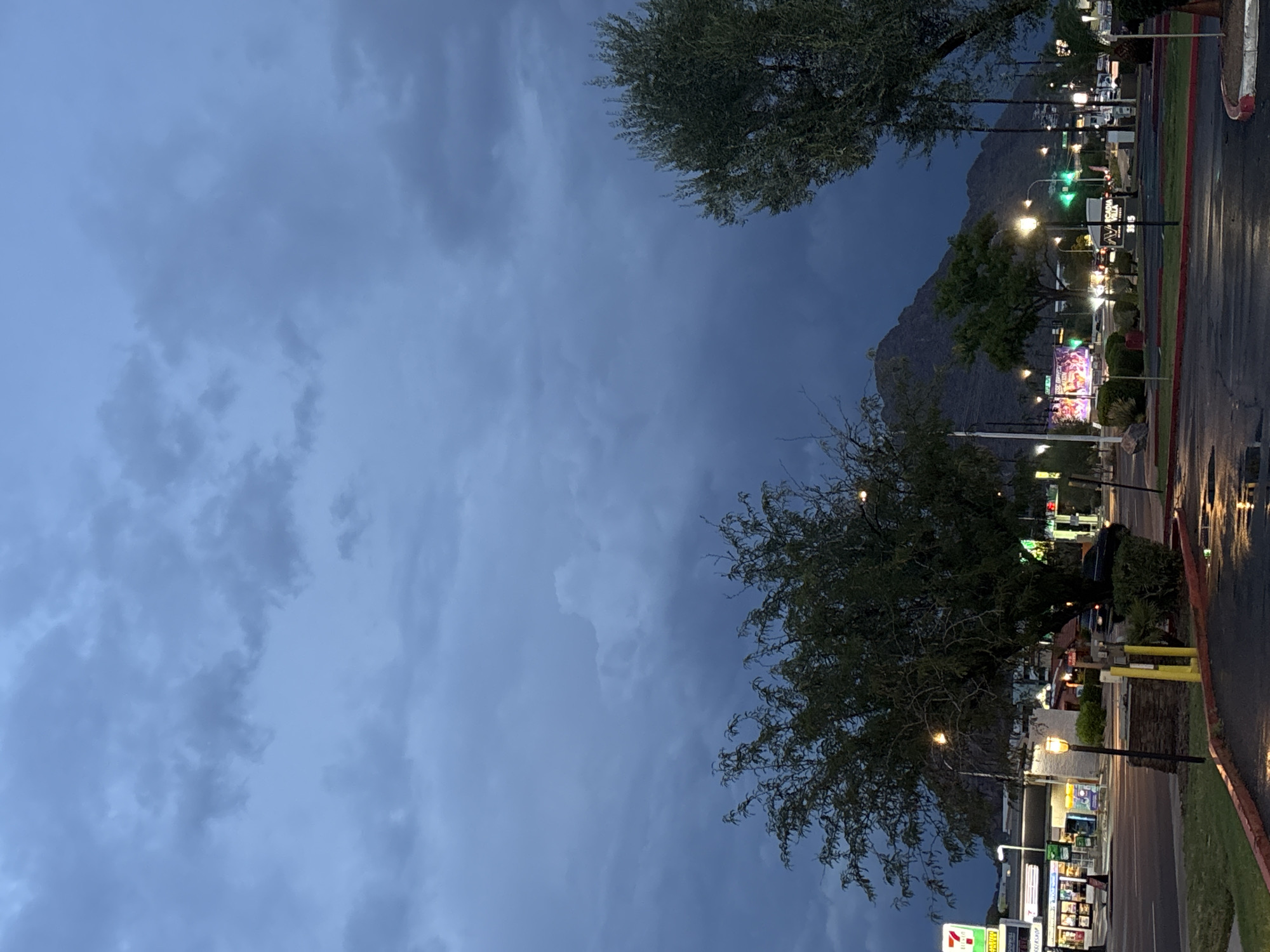 The image shows a cloudy sky over a city street with mountains in the background. The street is wet, suggesting it has recently rained. There are buildings on the left side of the street, including a 7-Eleven convenience store. A few cars are visible on the street, and there are streetlights and signs. There are trees on both sides of the street. The overall impression is of a somewhat gloomy, overcast day in an urban setting.