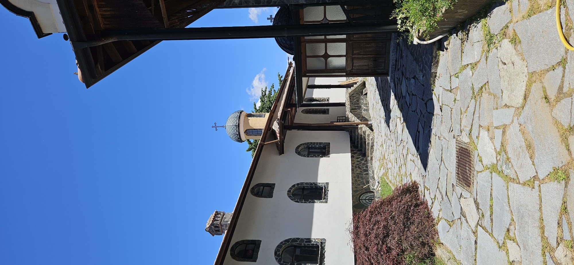 The image depicts a sunlit courtyard of a religious building, likely a church or monastery, in Ovchartsi, Bulgaria. The architecture features white walls with stone accents around arched windows. A small dome with a cross tops one of the structures, while another domed structure with a cross is partially visible on the right. The courtyard is paved with irregular grey flagstones, with grass growing in the gaps. A dark red Japanese maple tree stands to the left, and lush green foliage is visible in the background and along the right side. The sky is a clear, vibrant blue. The scene conveys a sense of peace and solitude under the bright midday sun.