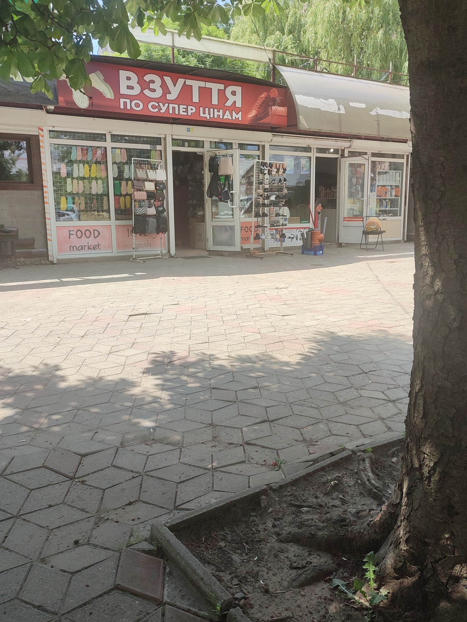 This is a full, eye-level, outdoor, daylight shot of a row of small market stalls or shops. The facade of the shops is visible, with a red awning above featuring white lettering that reads "ВЗУТТЯ ПО СУПЕР ЦІНАМ" (SHOES AT SUPER PRICES) and an image of a shoe and handbag. The stalls themselves have glass fronts showcasing merchandise. To the left, a stall displays rows of colorful flip-flops and sandals. Next to it, a stall features handbags and wallets. Further to the right, another stall has more shoes and bags on display, with an open doorway revealing more items inside. The stall to the far right has shelves stocked with various goods. The ground in front of the stalls is paved with hexagonal concrete tiles. A large tree trunk is visible on the right side of the frame, obscuring part of the scene. The background shows lush green foliage and trees. The general setting appears to be a pedestrian walkway or market area in Chernivtsi, Ukraine, likely on a sunny day.
