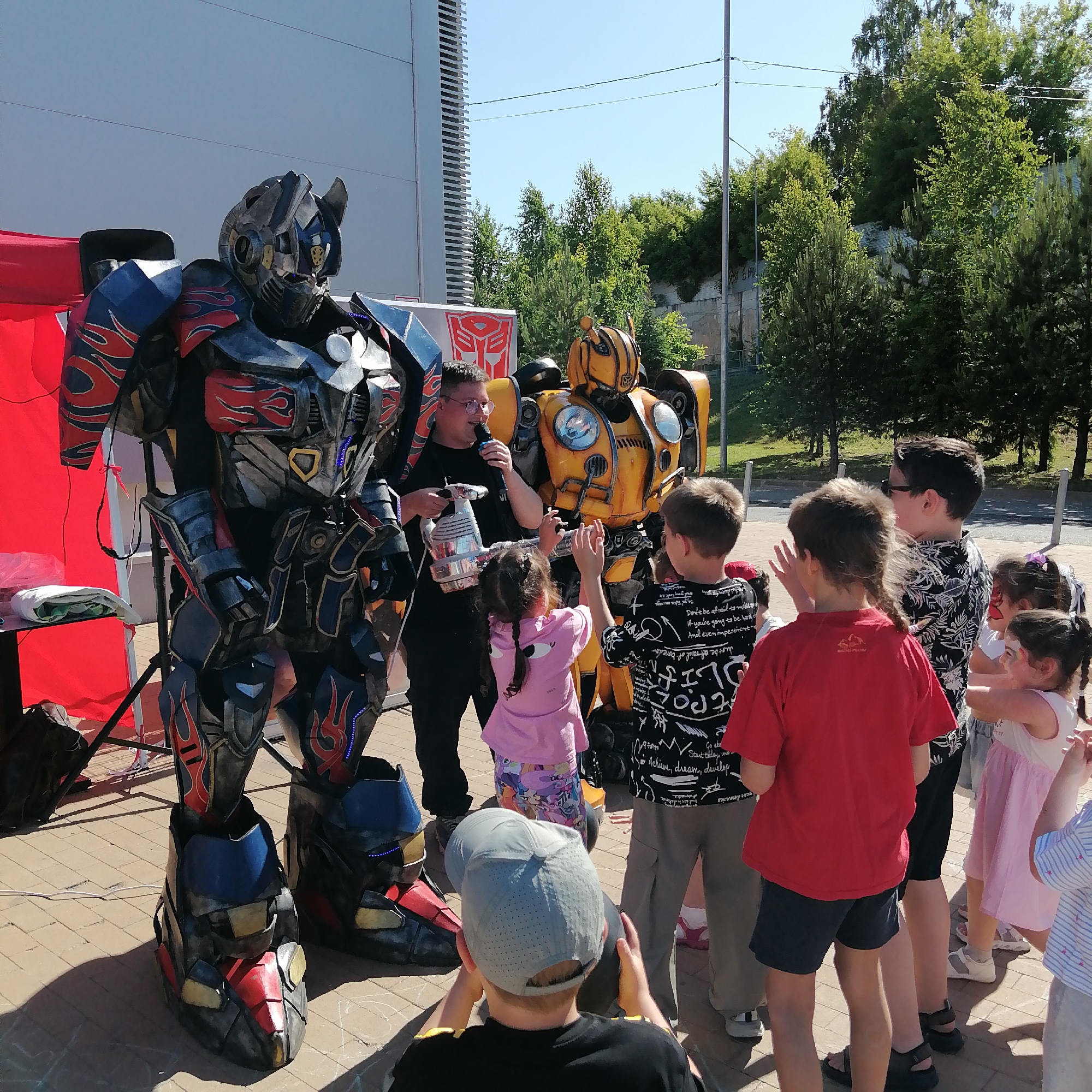 An outdoor event is depicted in Трубачеевка, Russia, under clear blue skies, featuring costumed performers and several children. In the foreground, a detailed, life-sized costume resembling Optimus Prime from the Transformers franchise stands prominently on a paved surface composed of light-colored pavers. This costume is predominantly dark gray and black, accented with red and blue armor panels, featuring stylized red flame patterns and illuminated blue lights on the chest and lower legs. A white electrical cord extends across the ground near its feet. To the right and slightly behind Optimus Prime, a person in dark attire, wearing glasses, holds a microphone to their mouth with their right hand and a silver, handheld device in their left. Adjacent to this individual, a second detailed costume, resembling Bumblebee, stands. This costume is primarily yellow with black accents and features visible headlights. Behind the performer and Bumblebee, a white banner or sign displays the red Autobot symbol.
A group of approximately eight children, ranging in age, are gathered around the performers. One girl with braided hair, wearing a pink shirt and patterned shorts, is extending her hands towards Bumblebee. Next to her, a boy in a black and white patterned shirt also has his hands raised towards Bumblebee. A boy in a red t-shirt and dark shorts stands with his back partially to the camera. Another boy, with his back completely to the camera, kneels or crouches on the ground, wearing a gray baseball cap and holding a yellow object. Further to the right, a girl with face paint (red and white design on her cheek) wears a light pink dress.
To the far left, a red canopy or tent structure is visible, with a small black table underneath holding a white folded cloth. The ground is paved with rectangular pavers, on which faint white chalk marks are visible. In the background, a large, light-colored building with distinct horizontal lines or panels rises on the left side. The background to the right consists of numerous green trees and dense foliage, with a utility pole and overhead power lines also present. A paved road or path is visible behind the children on the right side of the frame.