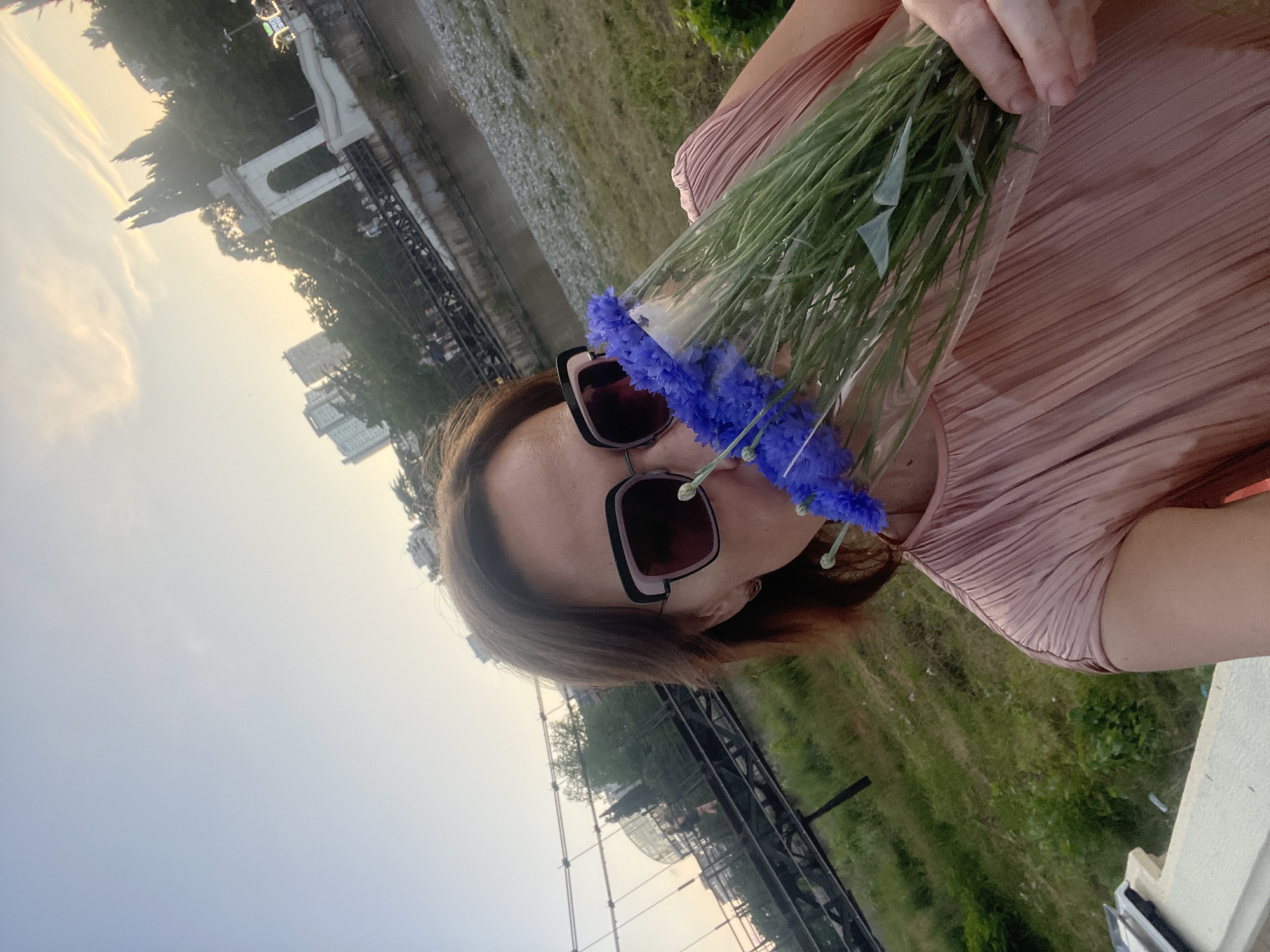 A woman wearing sunglasses and a pink pleated dress holds a bouquet of blue flowers, smelling them. She stands on a bank of grass and gravel, with a river and a suspension bridge in the background. Tall buildings and trees are visible on the other side of the river. A white archway with decorative spires is situated on the bridge. The scene appears to be in Sochi, Russia.