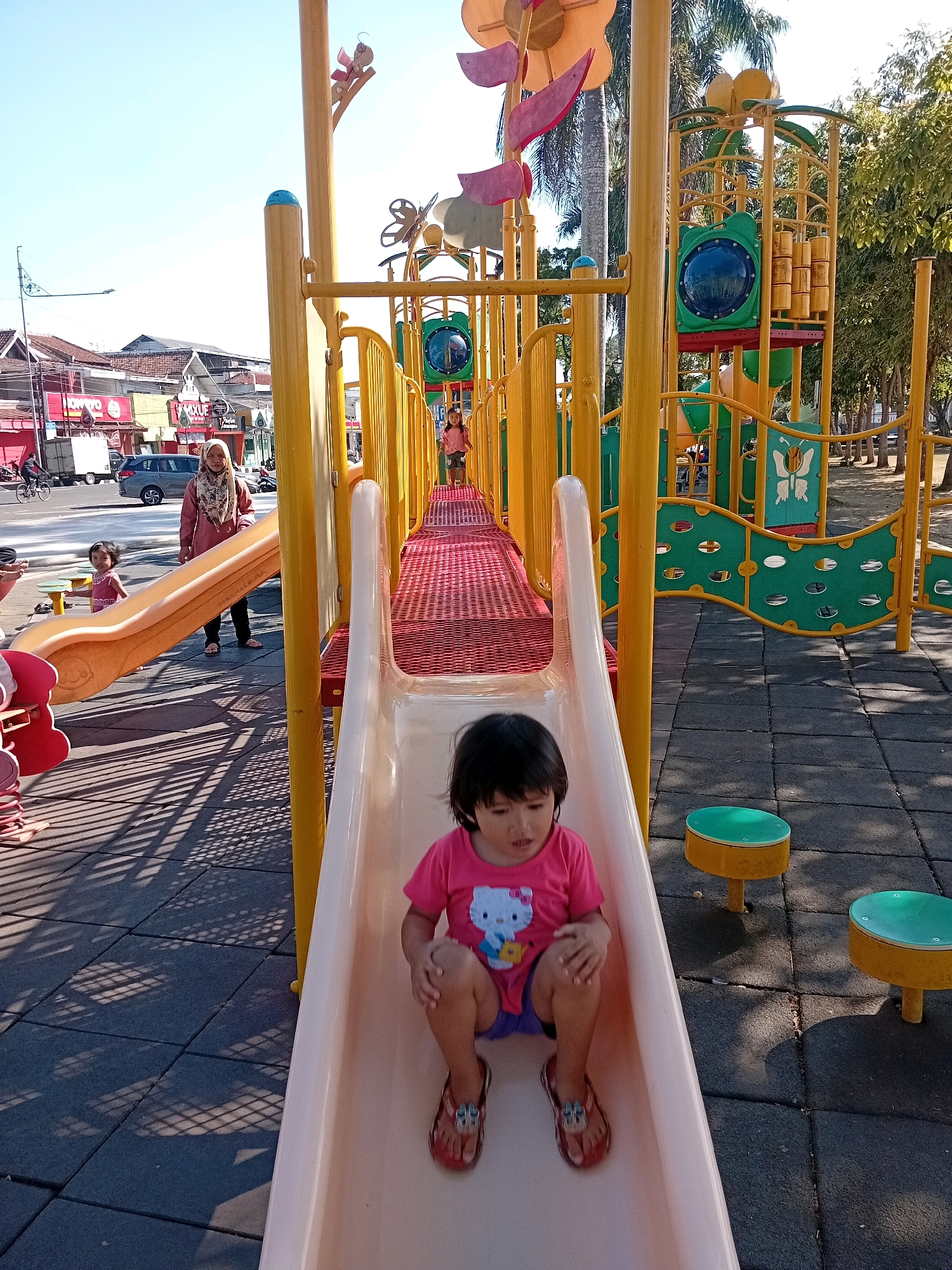 In Purworejo, Indonesia, a child with dark hair wears a pink t-shirt featuring Hello Kitty and blue shorts. They sit on a cream-colored slide, with their feet in red sandals touching the slide's surface. The child is positioned on the lower half of the slide. Behind the child, a red-meshed ramp leads upwards to a yellow playground structure. Further back, a woman in a peach-colored headscarf and outfit stands near the base of the slide. To the left of the woman, another young child in a pink shirt stands on a yellow and orange slide. The playground is situated on a dark gray rubberized surface with shadows cast from the playground structure. In the background, buildings line a street, and trees are visible. The sky is clear and blue.