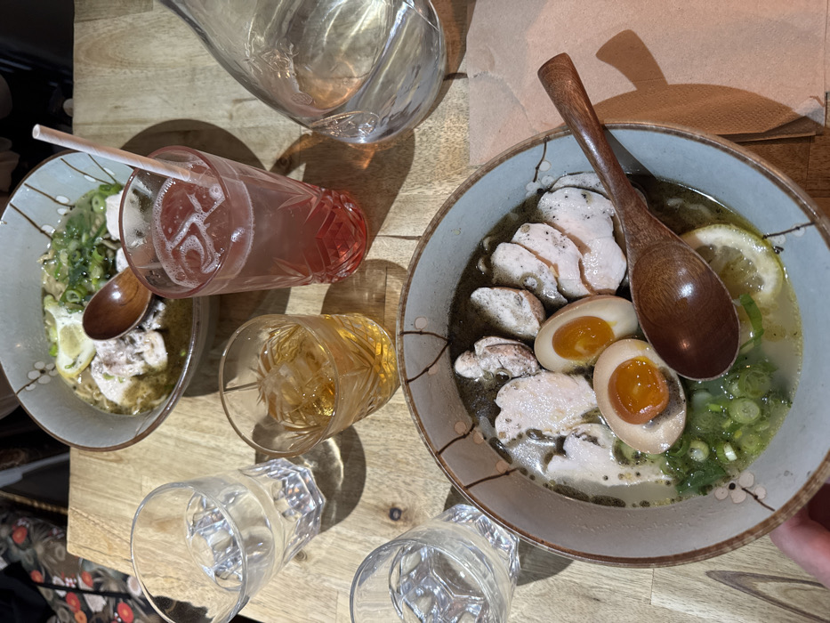 The scene depicts a light-colored wooden table set with food and beverages within an establishment in Paris, France. Two ceramic bowls of ramen are visible. The bowl in the foreground, held partially by a hand, contains ramen with a dark broth, slices of cooked white meat, two halved soft-boiled eggs with bright orange yolks, chopped green onions, and a lemon slice, accompanied by a dark wooden spoon resting on its rim. The background bowl contains ramen with a lighter, greenish broth, slices of cooked white meat, chopped green onions, and a lemon wedge, with a small wooden spoon submerged. Beverages on the table include a tall, textured glass holding a reddish-pink iced drink with a white paper straw, a shorter, textured glass containing an amber iced drink, two clear faceted glasses filled with water, and a glass carafe of water. Shadows are cast across the wooden table surface.