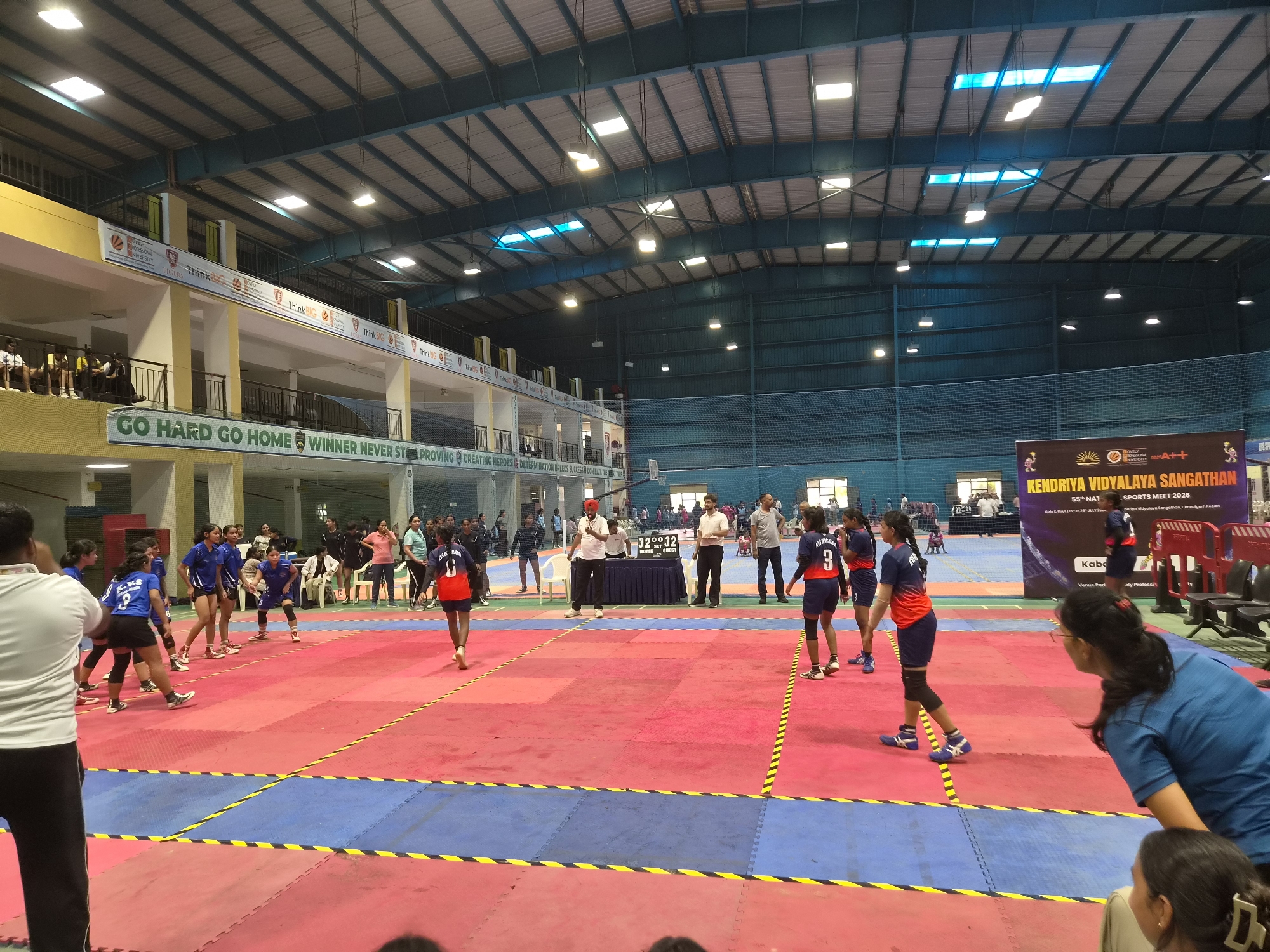 The image shows an indoor sports arena, likely a gymnasium, with a bright red and blue mat laid out for a game. Several young women are actively playing on the mat, dressed in blue and red sports uniforms. Some are in mid-action, while others appear to be observing. Spectators are visible in the background, some seated in bleachers, and others standing. There are banners and signage around the arena, indicating a sports meet. One prominent banner reads "KENDRIYA VIDYALAYA SANGATHAN SPORTS MEET 2026". The architecture of the building, with its high ceiling and metal beams, suggests a large public indoor facility. The lighting appears to be a mix of fluorescent and natural light from skylights. Based on the provided information, this scene is located in Phagwara, India, within the Kendriya Vidyalaya Sangathan Sports Meet.