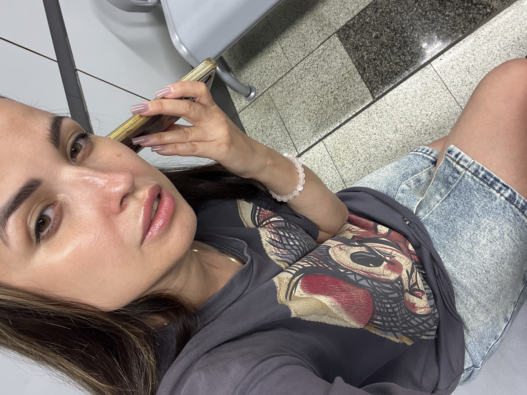 A woman is holding a smartphone to her ear, implying she is on a call. She is wearing a dark grey t-shirt with a graphic print and light blue denim shorts. A pale pink beaded bracelet is visible on her left wrist. She has long, dark brown hair with lighter highlights. The background appears to be an indoor waiting area, possibly a public transport station, with tiled flooring and grey bench seating. The lighting is bright, possibly from overhead fixtures, and casts highlights on her skin.