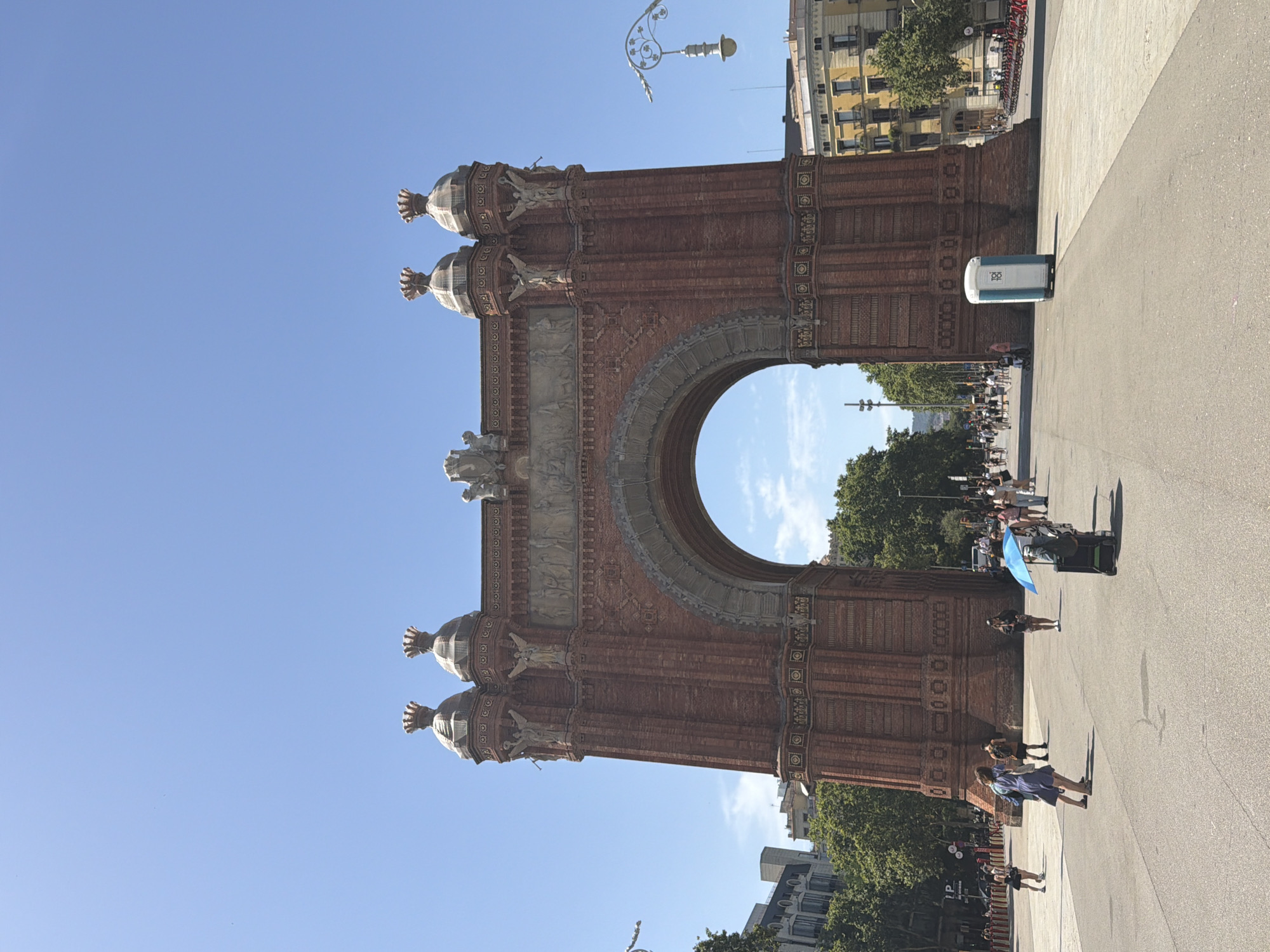 The Arc de Triomf in Barcelona, Spain, is captured from a low angle on a clear, sunny day. The monument, constructed of red brick and adorned with sculptures, stands prominently against a bright blue sky. Several people are visible in the foreground, walking on a paved plaza. A portable toilet is situated to the right of the arc, and in the background, trees and buildings line the street.