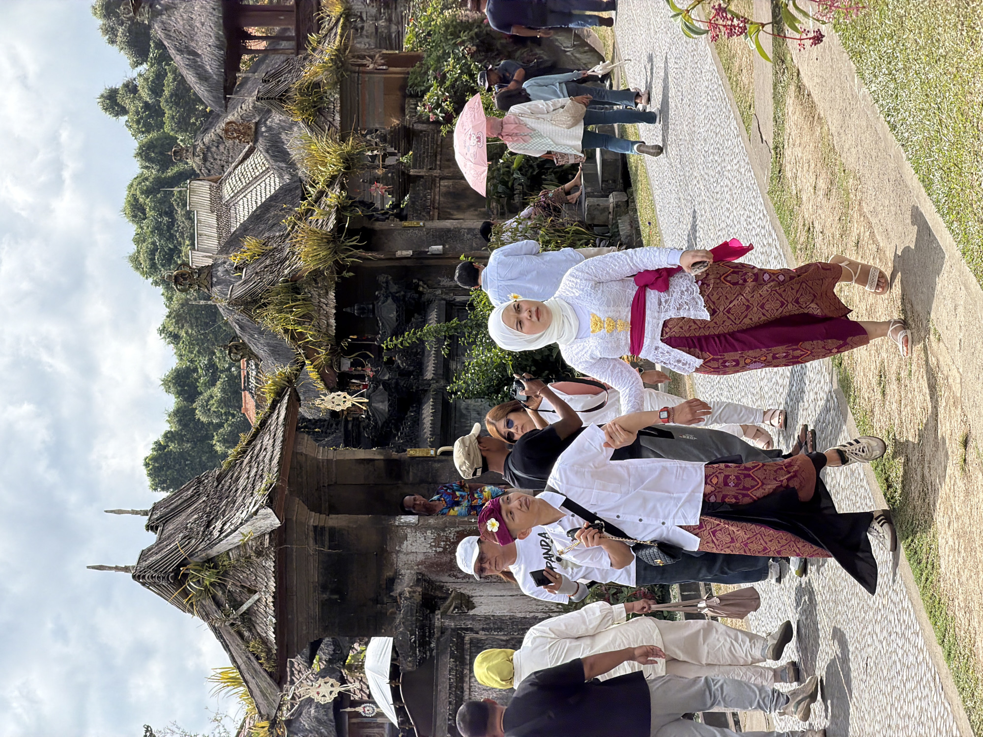 In Susut, Indonesia, a group of tourists and locals walk along a paved path in front of traditional Balinese buildings with thatched roofs and ornate carvings. In the foreground, a couple dressed in traditional attire walks together, the man in a white shirt and patterned sarong, and the woman in a white lace top and matching sarong, both wearing headscarves. The man is holding the woman's hand. Other individuals are seen in the background, some holding cameras and umbrellas, suggesting a cultural or tourist site. Lush greenery and trees frame the scene, indicating a tropical environment. The sky is partly cloudy.