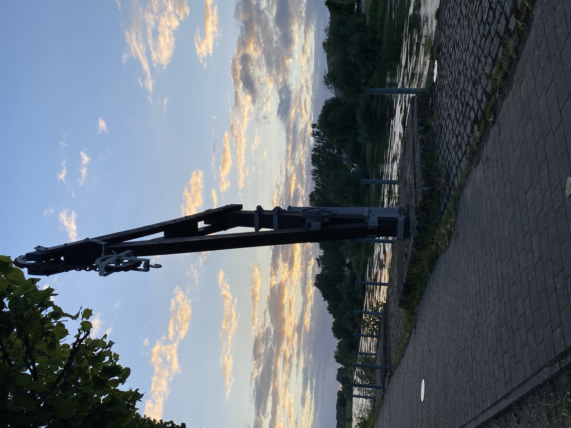 The image shows a view of the Weser River in Rinteln, Germany, at sunset. In the foreground, a paved walkway lines the riverbank. To the right of the walkway is a dark, wooden structure with metal accents, possibly a historical winch or crane, with a chain and hook visible. The river reflects the warm colors of the sunset sky, which is filled with scattered, light-colored clouds tinged with orange and pink. Dense green trees form the opposite bank of the river. A metal fence runs along the river's edge. The scene is tranquil, with no people or animals visible.