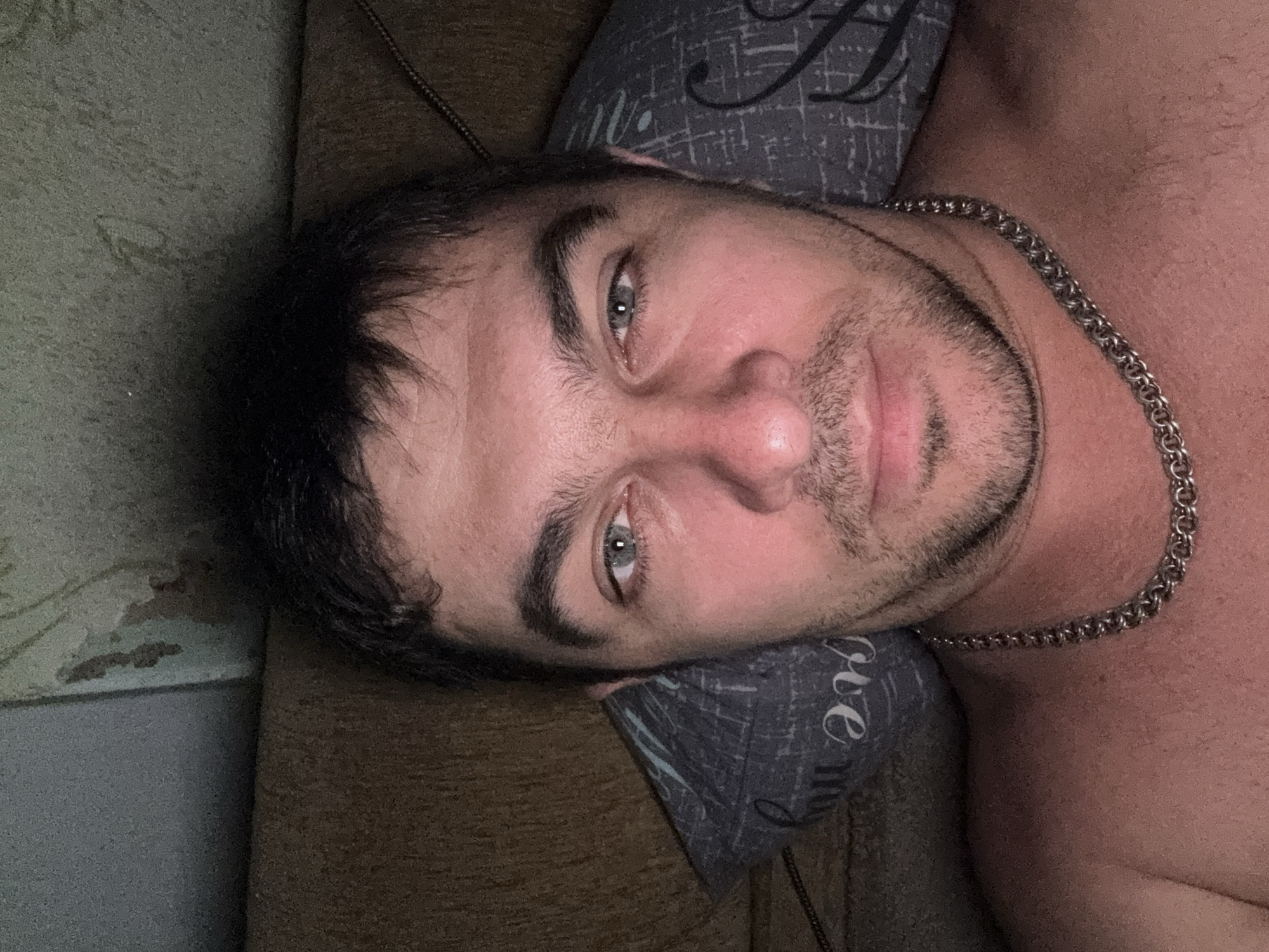 The image shows a close-up selfie of a man lying down, with his head resting on a pillow. He has short, dark hair, prominent eyebrows, and light-colored eyes. He is wearing a silver chain necklace. The background appears to be a couch and a wall with some textured wallpaper. The lighting suggests an indoor setting, possibly at night. There is no visual information within the image to determine the specific location in Shakhty, Russia.
