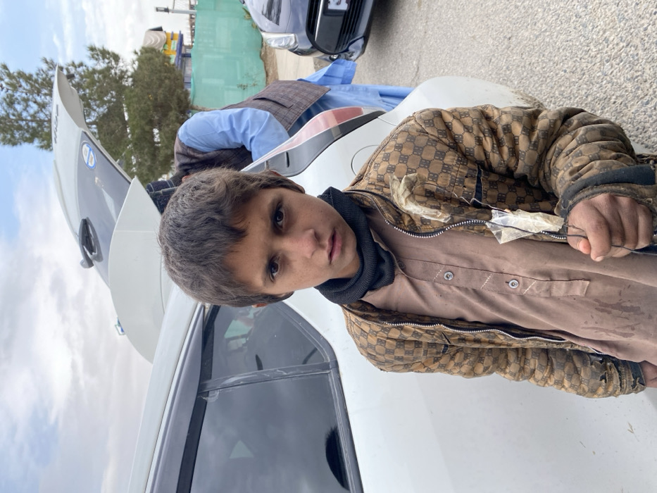 A young male, estimated between 8 to 12 years old, stands centrally in the foreground, directly facing the observer. He has short, dark, disheveled hair, and his face shows dirt smudges. He is wearing a black neck gaiter, a brown long-sleeved collared shirt with buttoned placket and white decorative stitching, and a dirty, torn, brownish-tan quilted jacket featuring a repetitive dark pattern. The jacket has visible damage, particularly on the left sleeve and near the zipper. In his right hand, he holds a thin, dark wire or string from which a small, clear plastic bag containing a light-colored, amorphous substance is suspended.
Behind the boy, a white multi-purpose vehicle (SUV or hatchback) is parked, with its rear hatch open, revealing the cargo area. To the right of the boy, behind the white vehicle, a male adult wearing a light blue shirt and a dark patterned vest is partially visible, appearing to lean towards or interact with the open trunk. A dark-colored sedan is partially visible to the far right. The environment is an outdoor, paved area, likely a street or parking lot, under an overcast sky. In the distant background, a large tree with sparse foliage and a green fence are present. The scene is located in Kandahar, Afghanistan.
